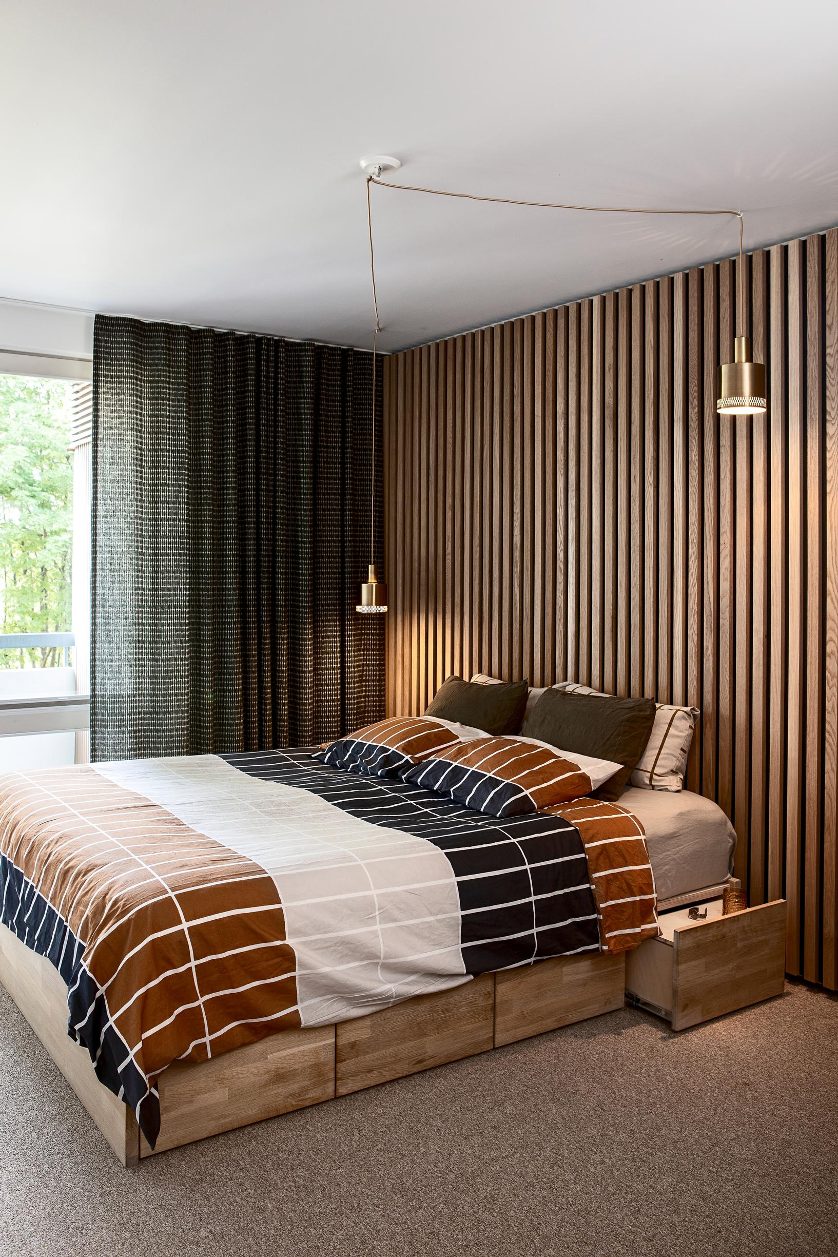 The headboard wall of the double bed is slatted. There is a dark curtain on the window wall. The duvet cover and pillowcases feature brown, black, and white.