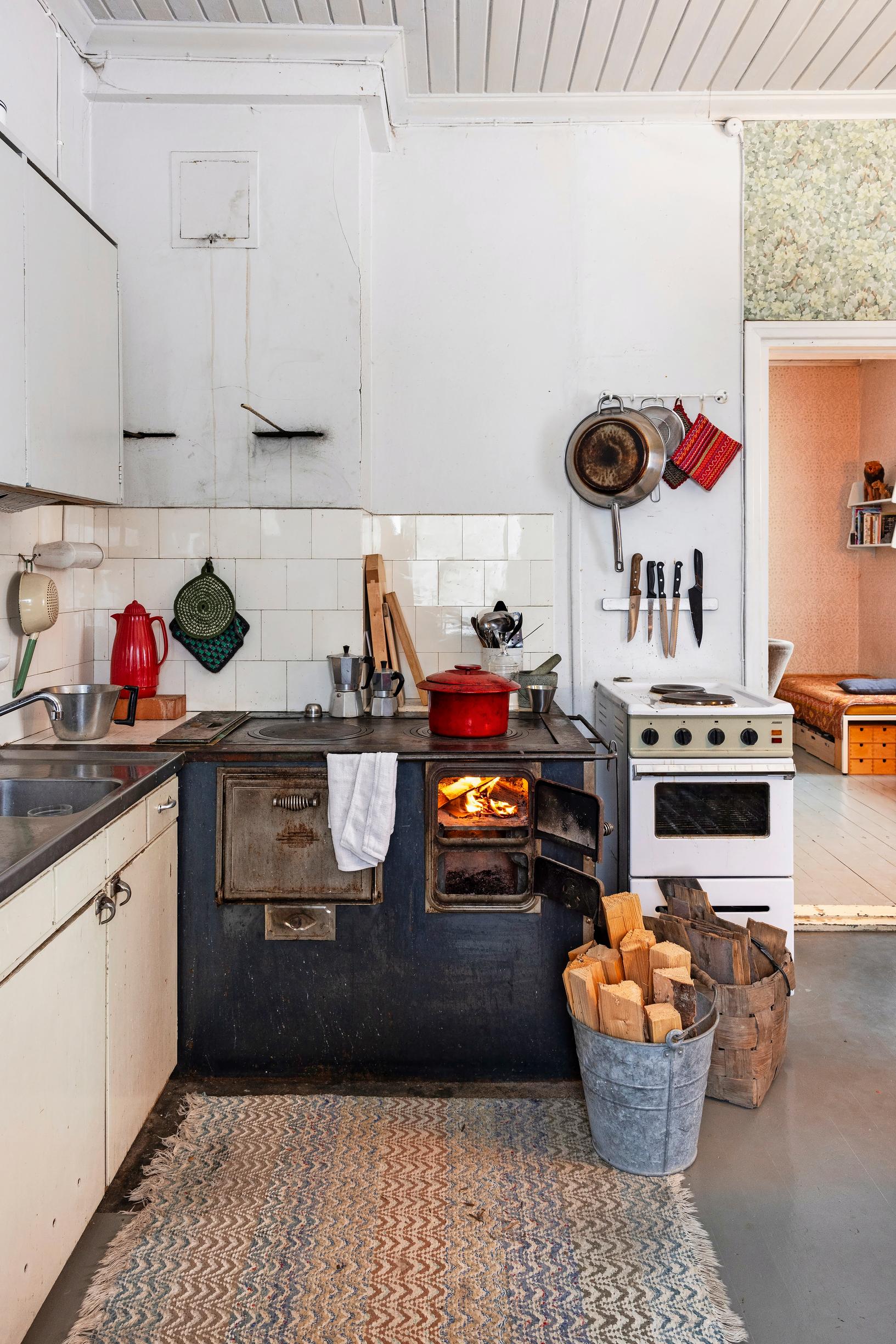 In a white kitchen, a dark wood stove with a fire, logs in a basket and in a bucket