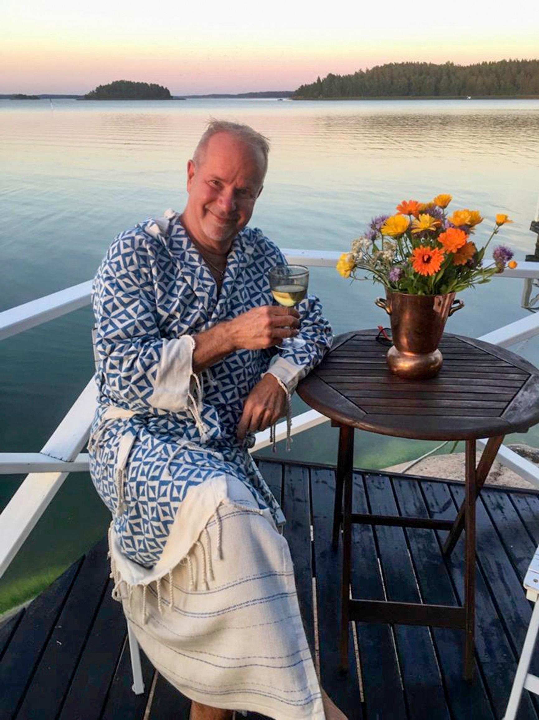 Singer Petri Laaksonen in a bathrobe on the sauna terrace, holding a glass. Marigolds on the table, with a sunset over the sea in the background