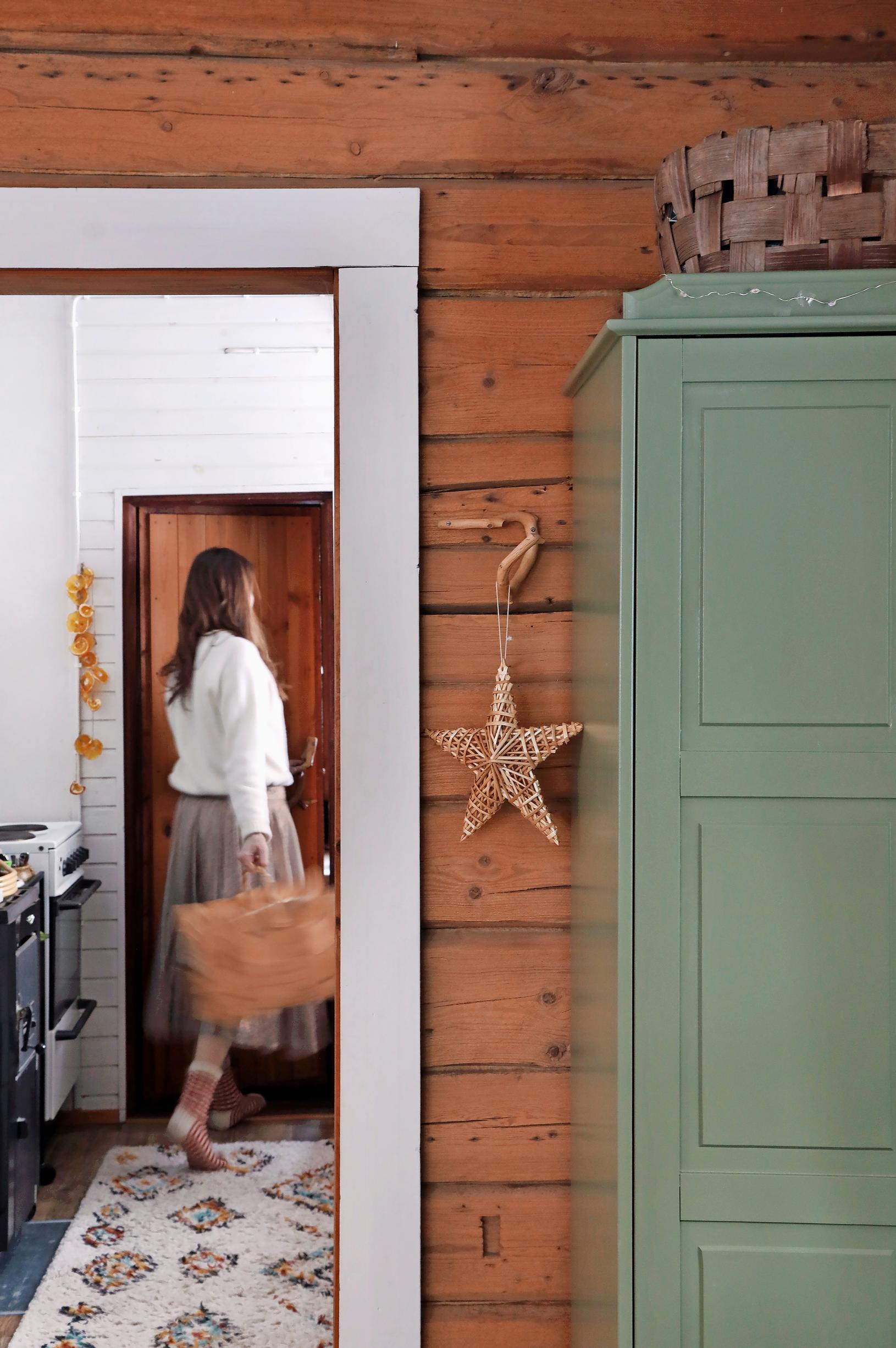 Ulla Parviainen stepping through the cabin door with a basket in hand, with a green cabinet in the foreground.