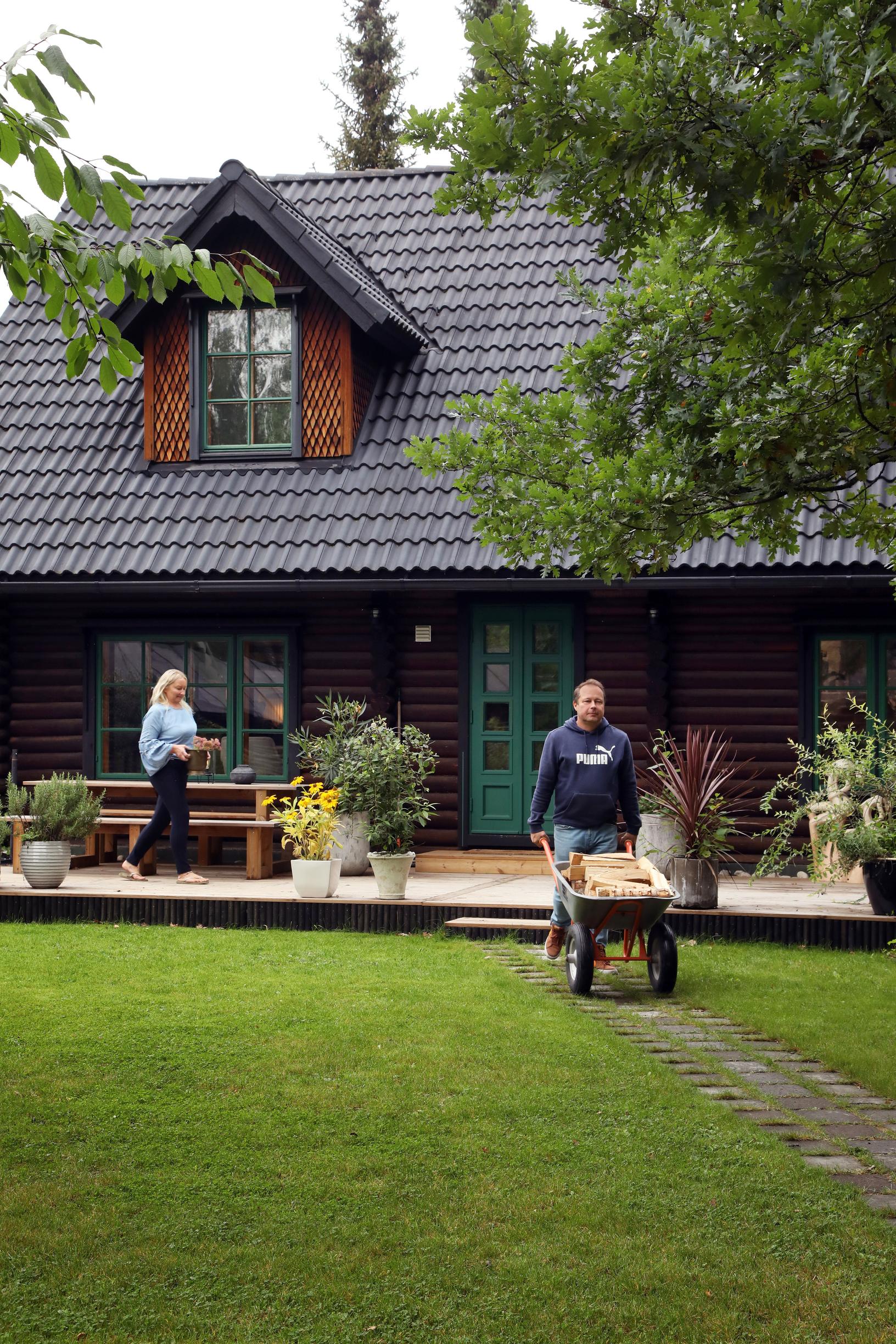 Katja stands on the log house deck while Kimmo pushes a wheelbarrow.