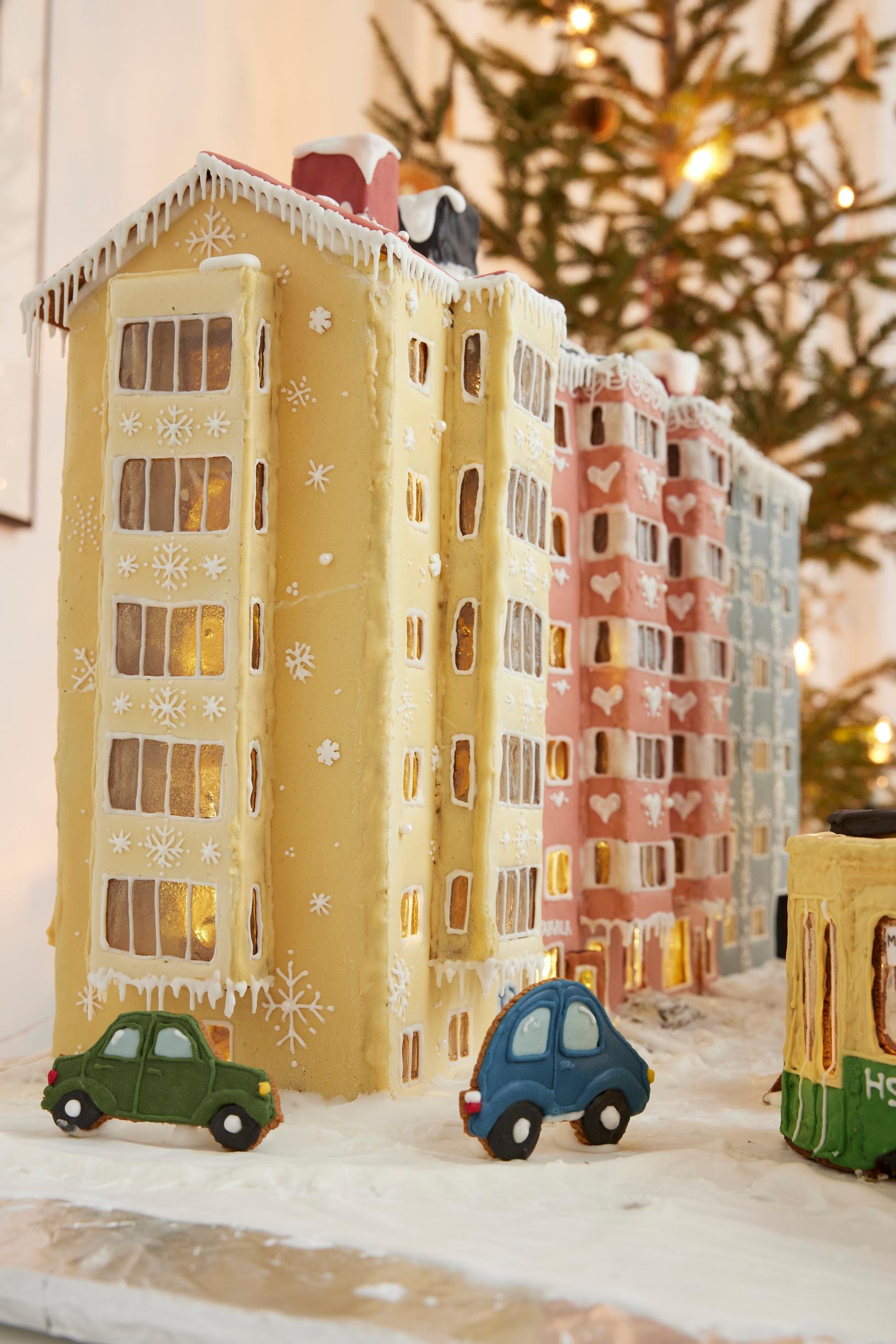 A gingerbread city block featuring a 1950s apartment building with bay windows covered in yellow and pink royal icing.