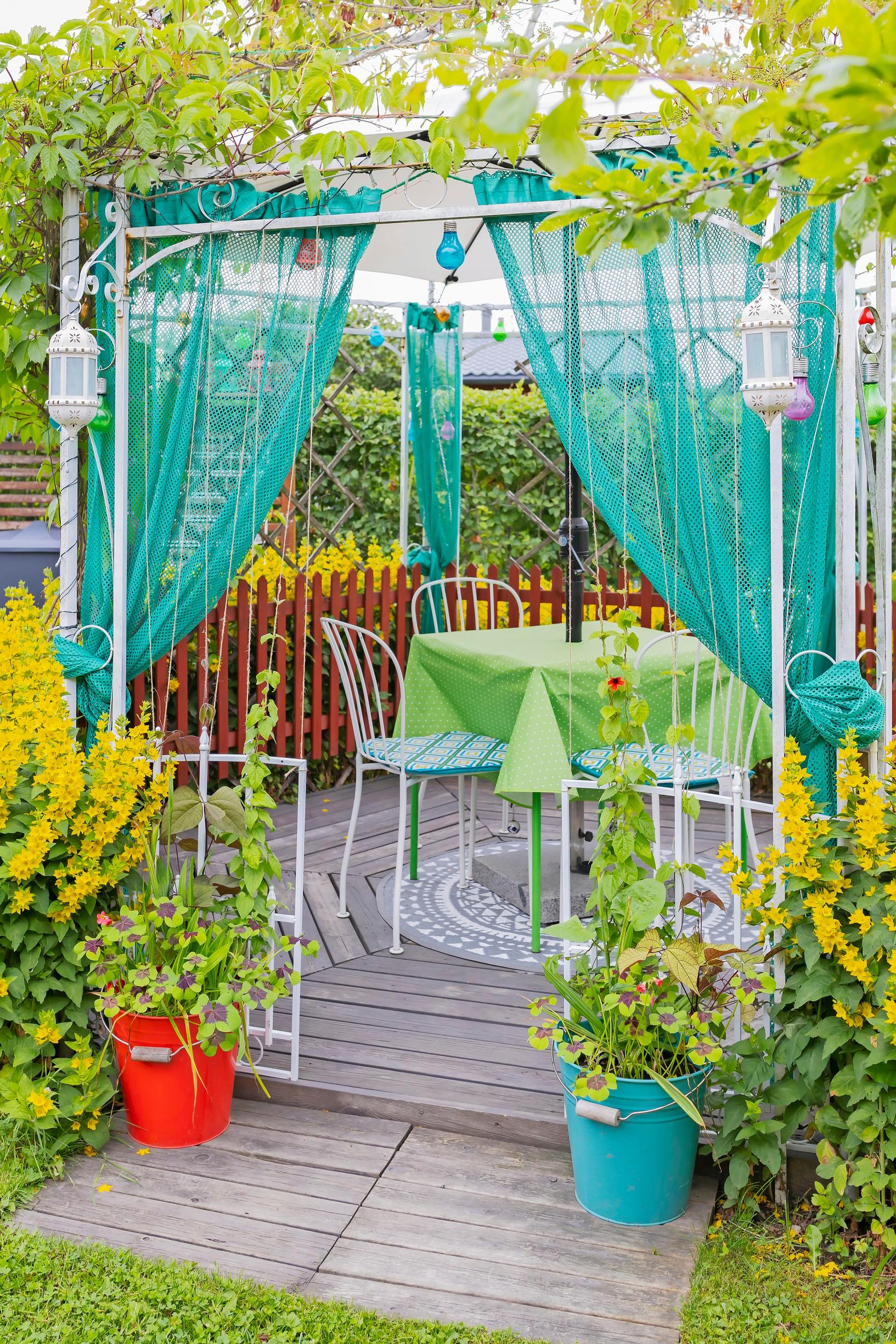 an outdoor canopy with green pleated curtains and a dining set, flowers in buckets at the door, yellow loosestrife