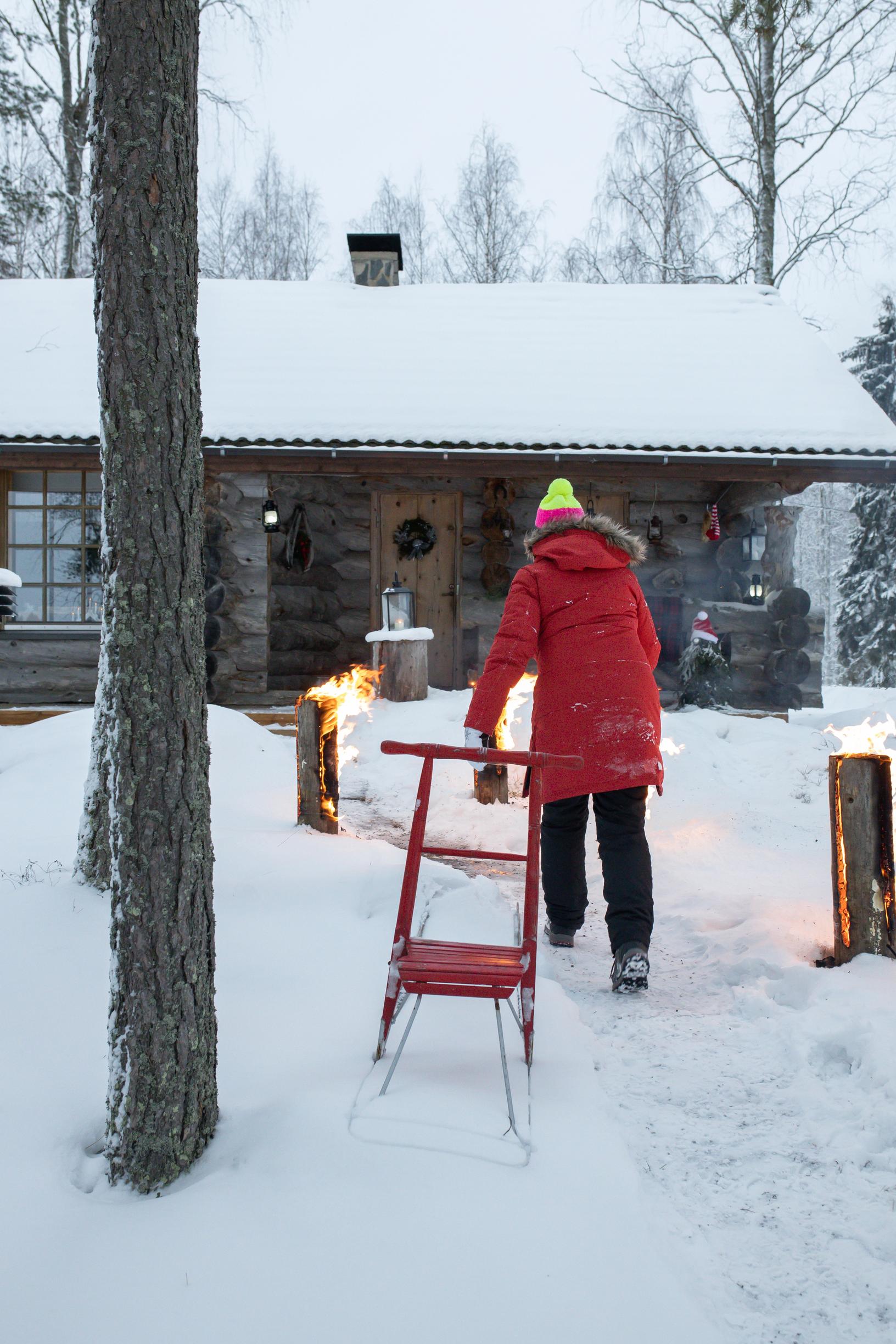 A woman walks toward the kelo cabin through a snowy yard, which has a red kicksled and burning log torches.