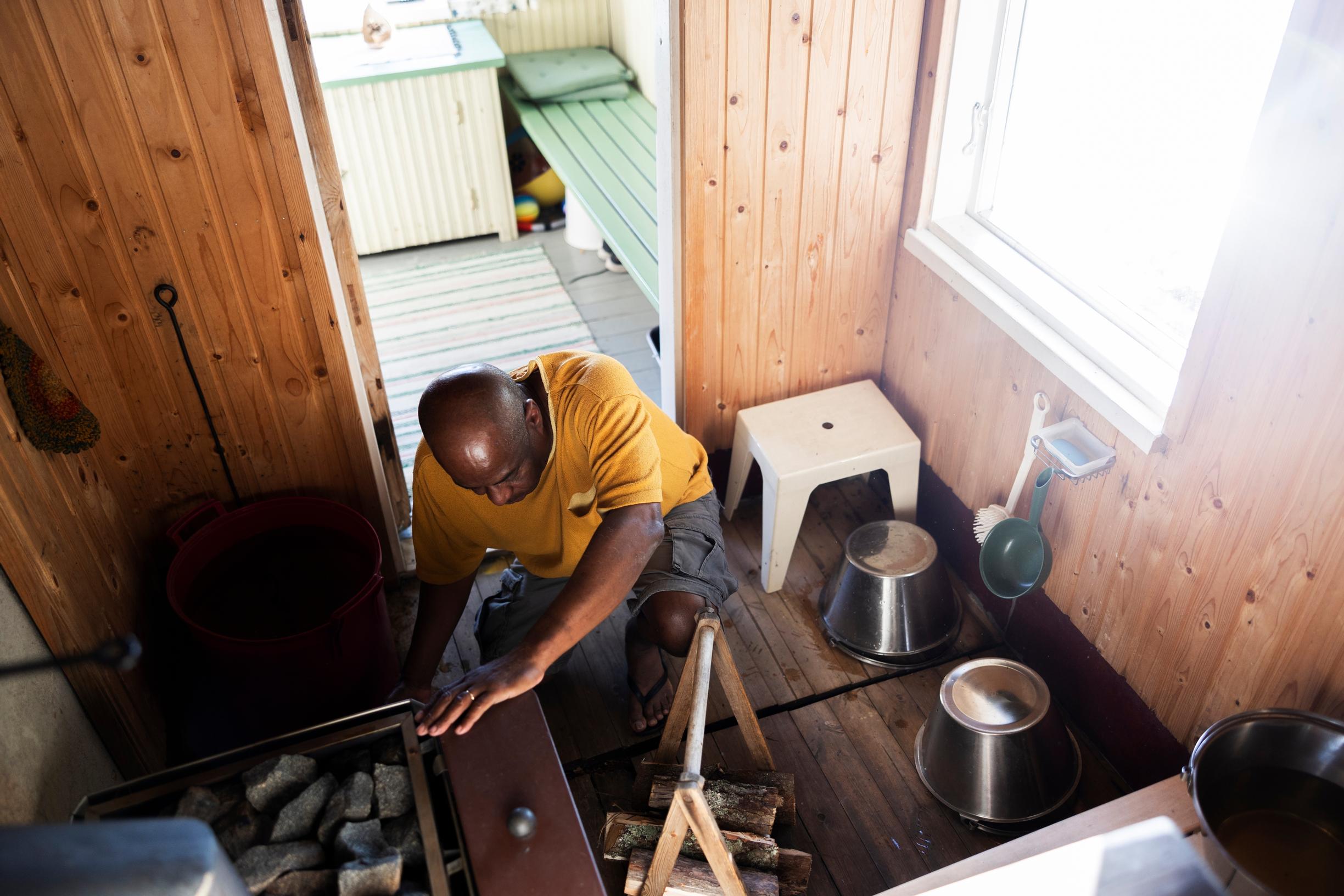 Edson placing wood into the sauna stove’s firebox, buckets in the sauna, view to the dressing room