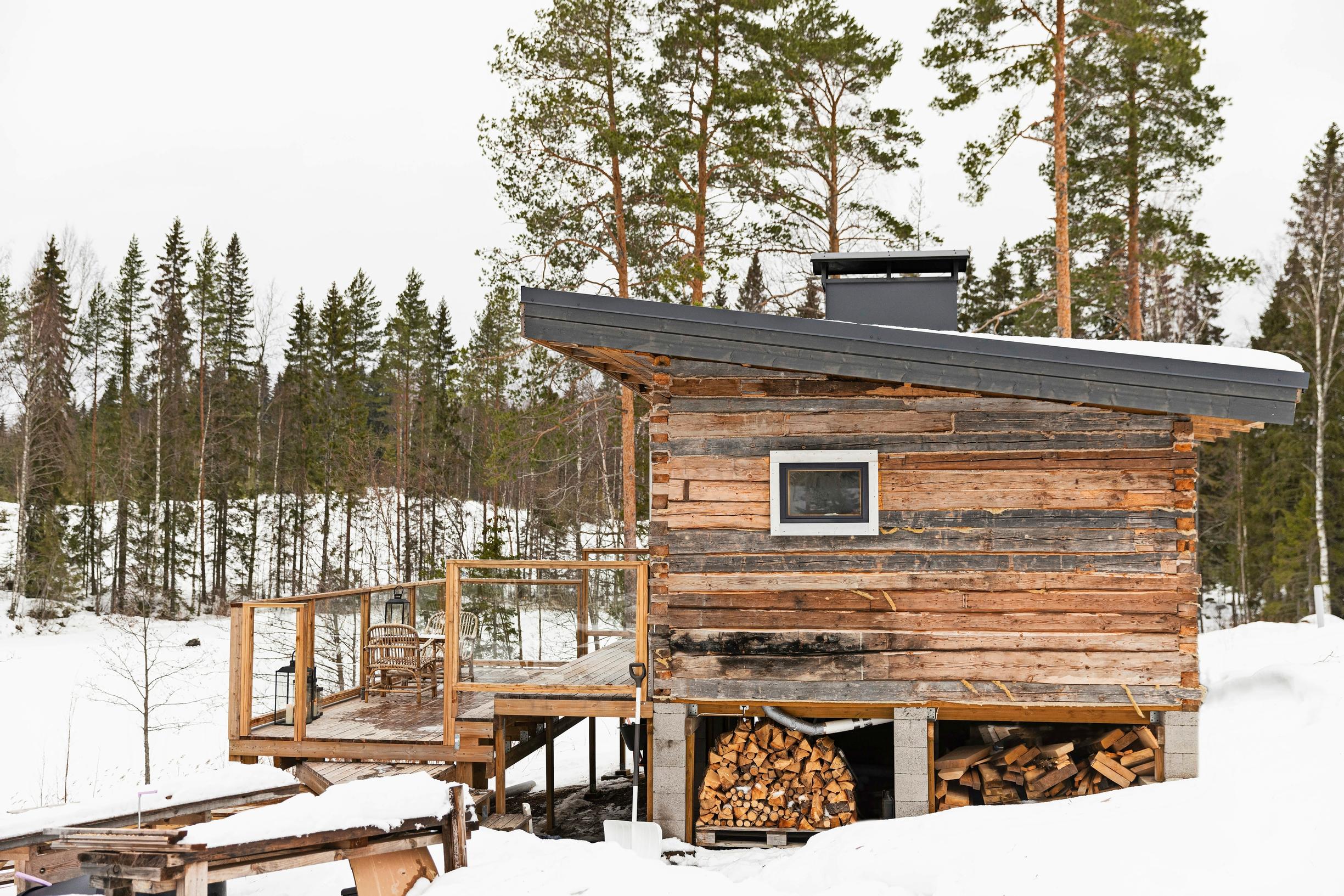 A weathered log building on a snowy lakeshore, with a forest in the background.