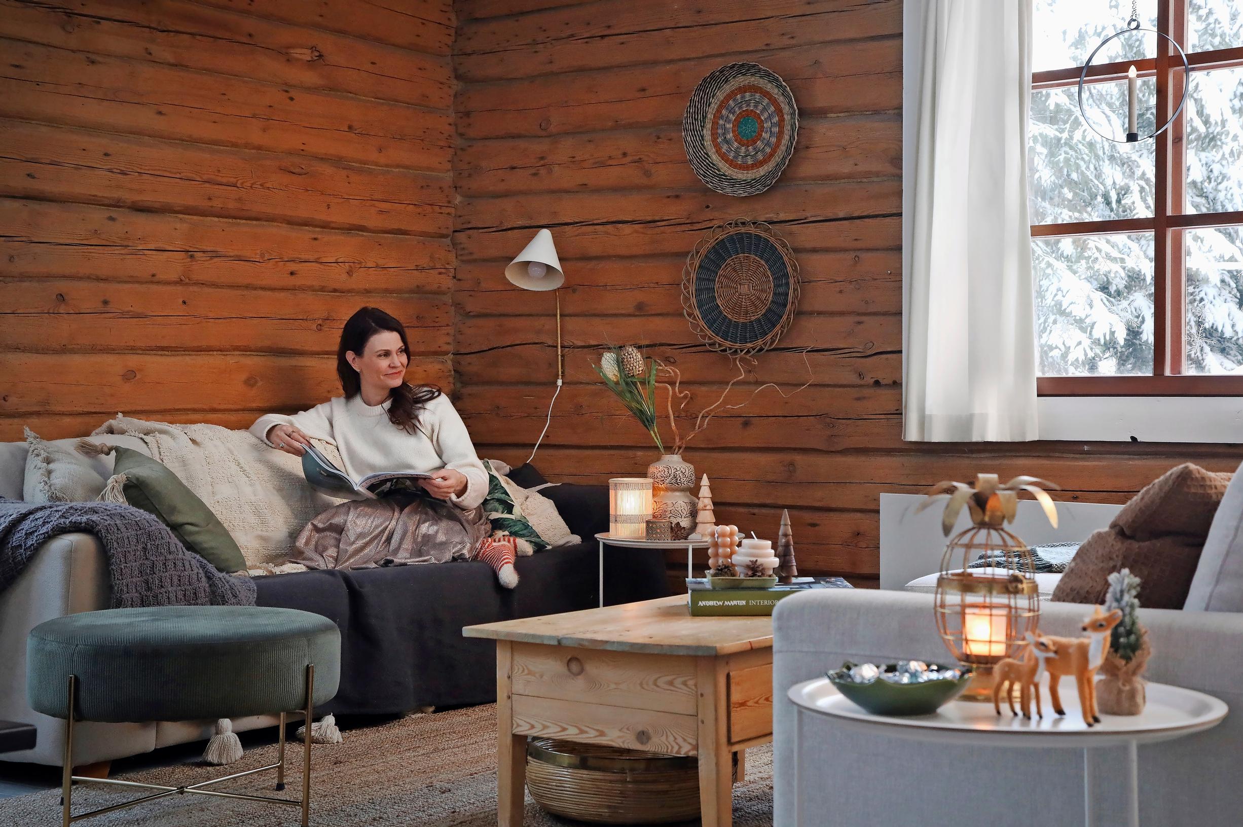 Ulla Parviainen sits on the sofa reading, surrounded by an ottoman, an armchair, a coffee table, and a few side tables, with a log wall and a forest view in the window.