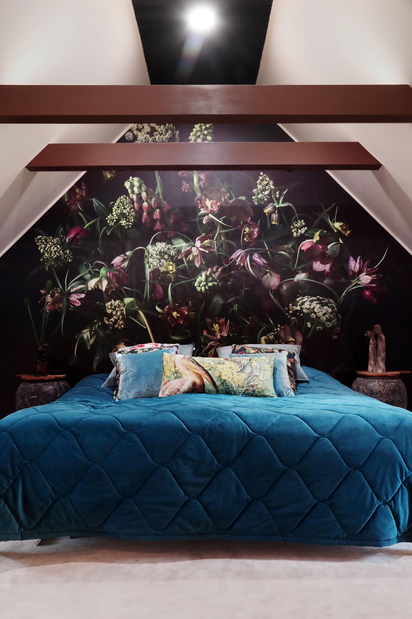 The bedroom has a sloped ceiling and exposed beams. There’s a dark patterned wallpaper behind the bed.