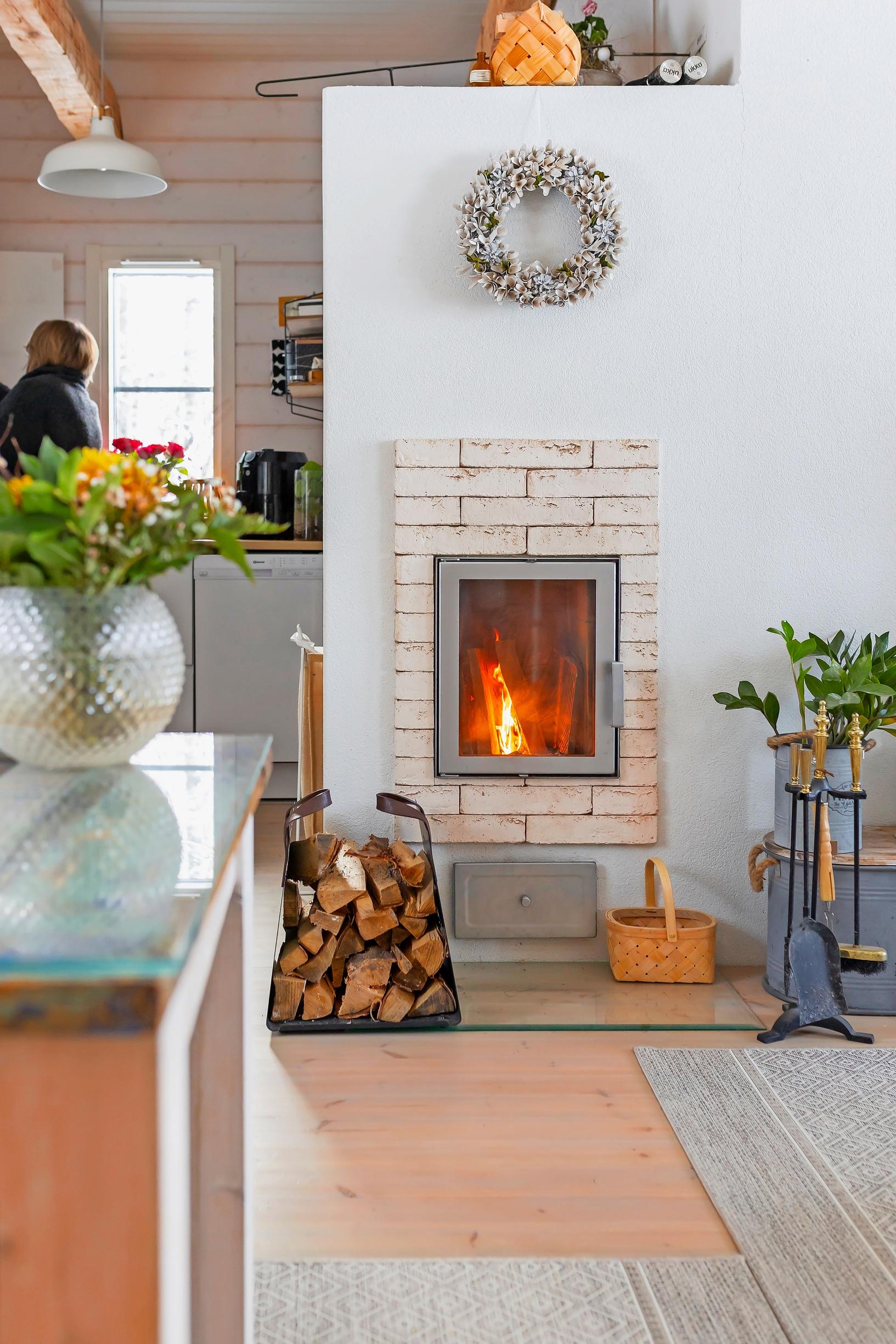 A light-colored brick fireplace with a fire burning, a log holder with wood in front, a woman in the kitchen in the background