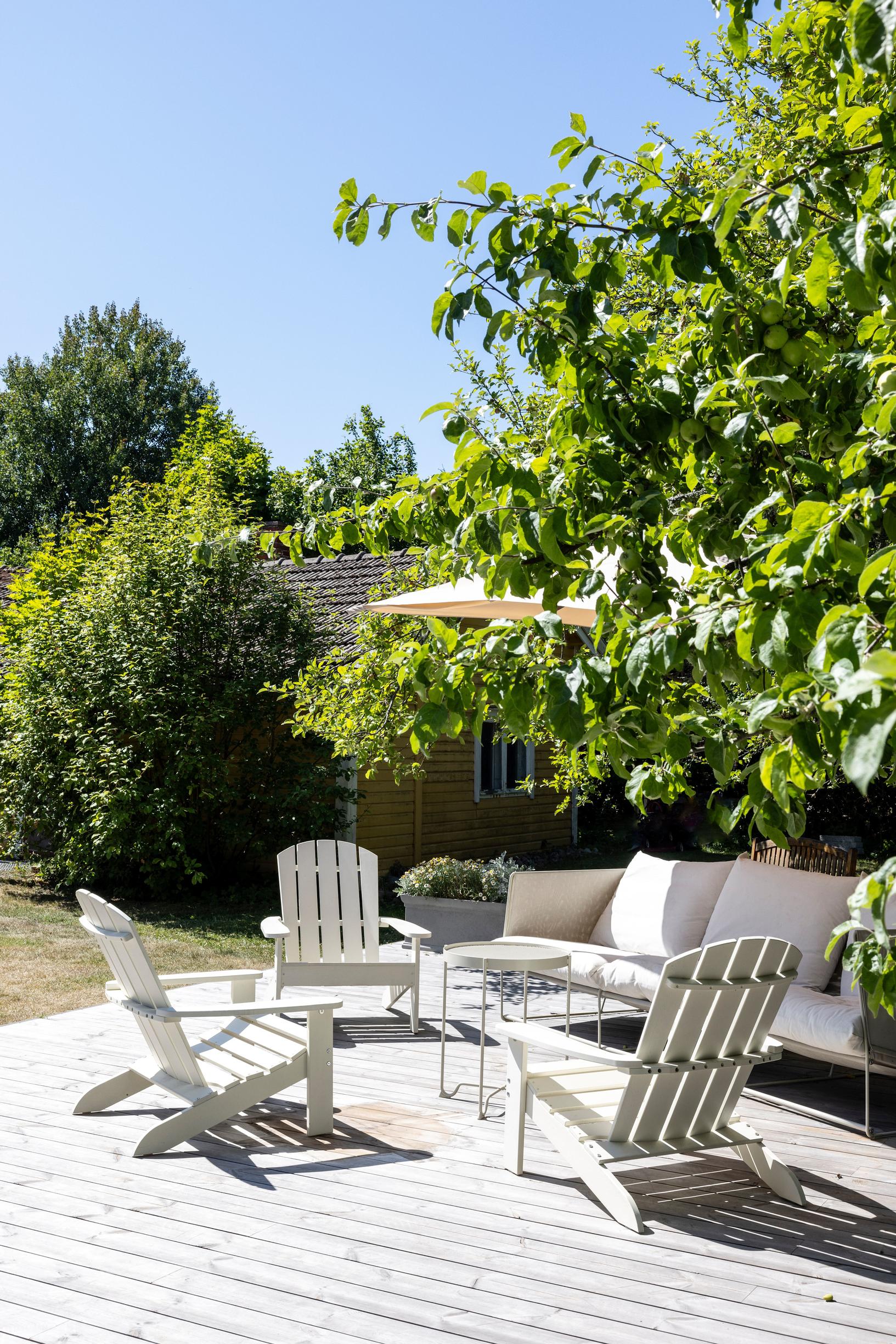 On the patio, there are white wooden outdoor furniture pieces surrounded by shrubs and trees.