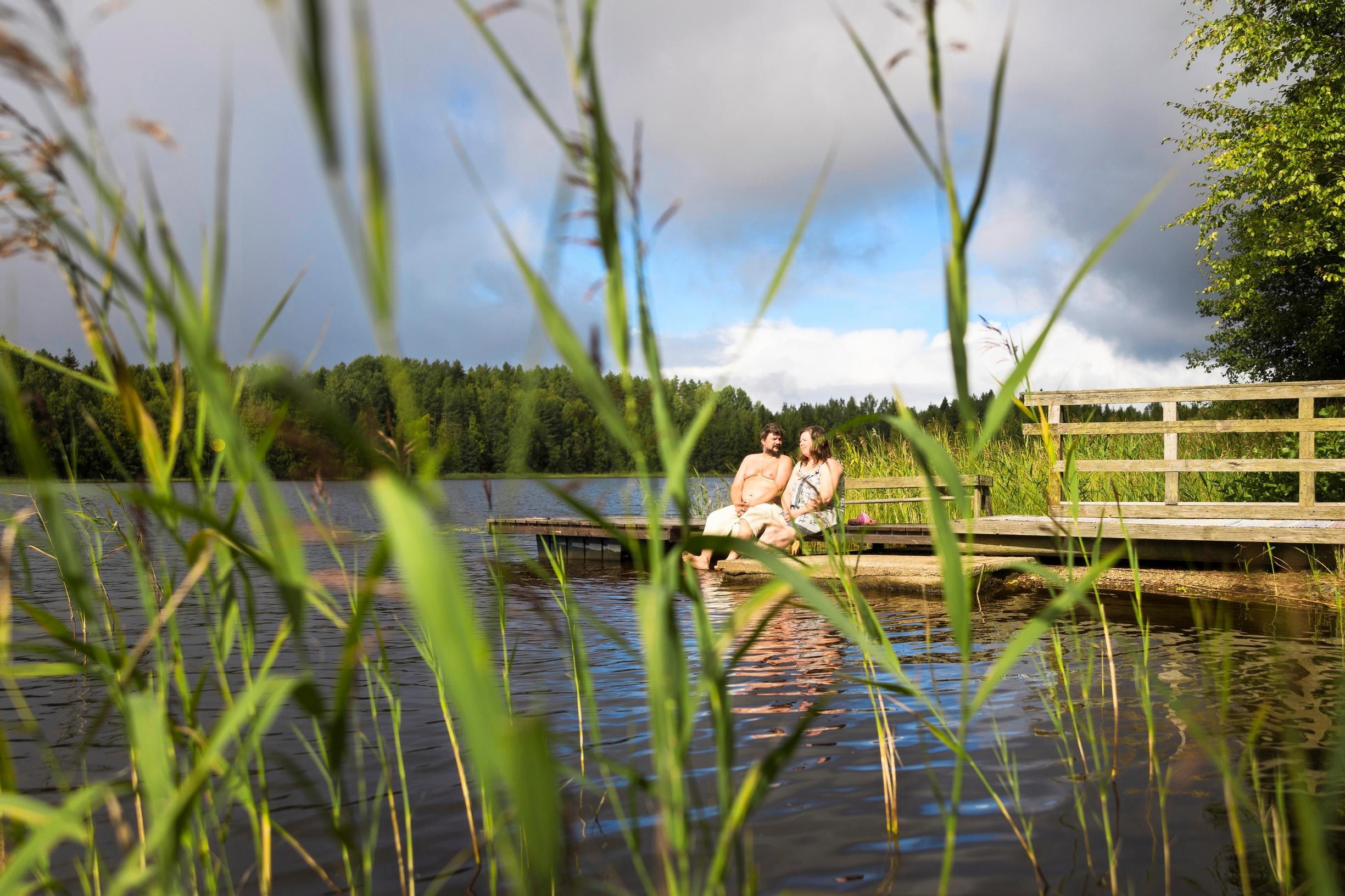 A woman and a man seated on the dock, looking at each other, with reeds in the foreground.