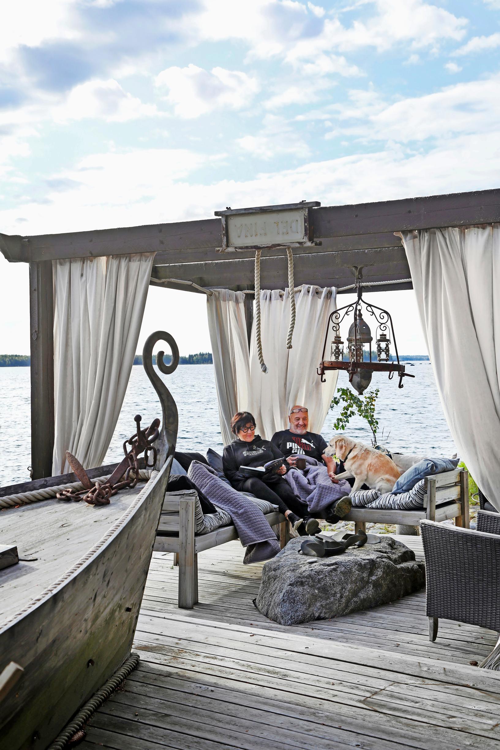 Sanna and Markku Parkkari sitting on a covered dock with their dog on a corner sofa, with the bow of a boat in the foreground.