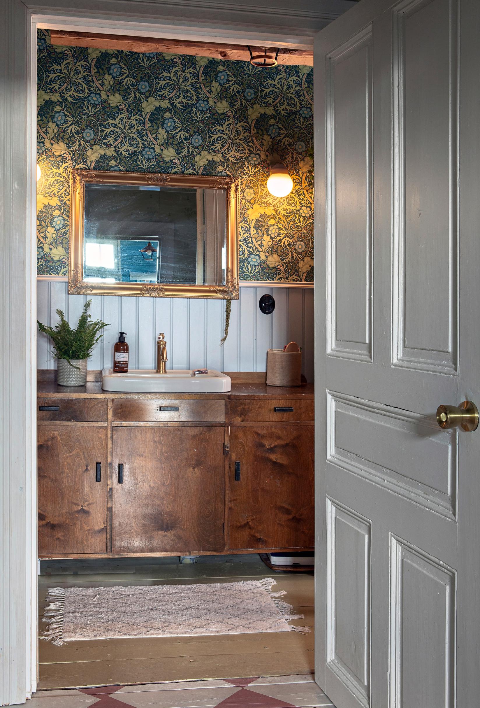 The renovated bathroom of the old house features a sink cabinet made from a chest of drawers and colorful William Morris wallpaper.