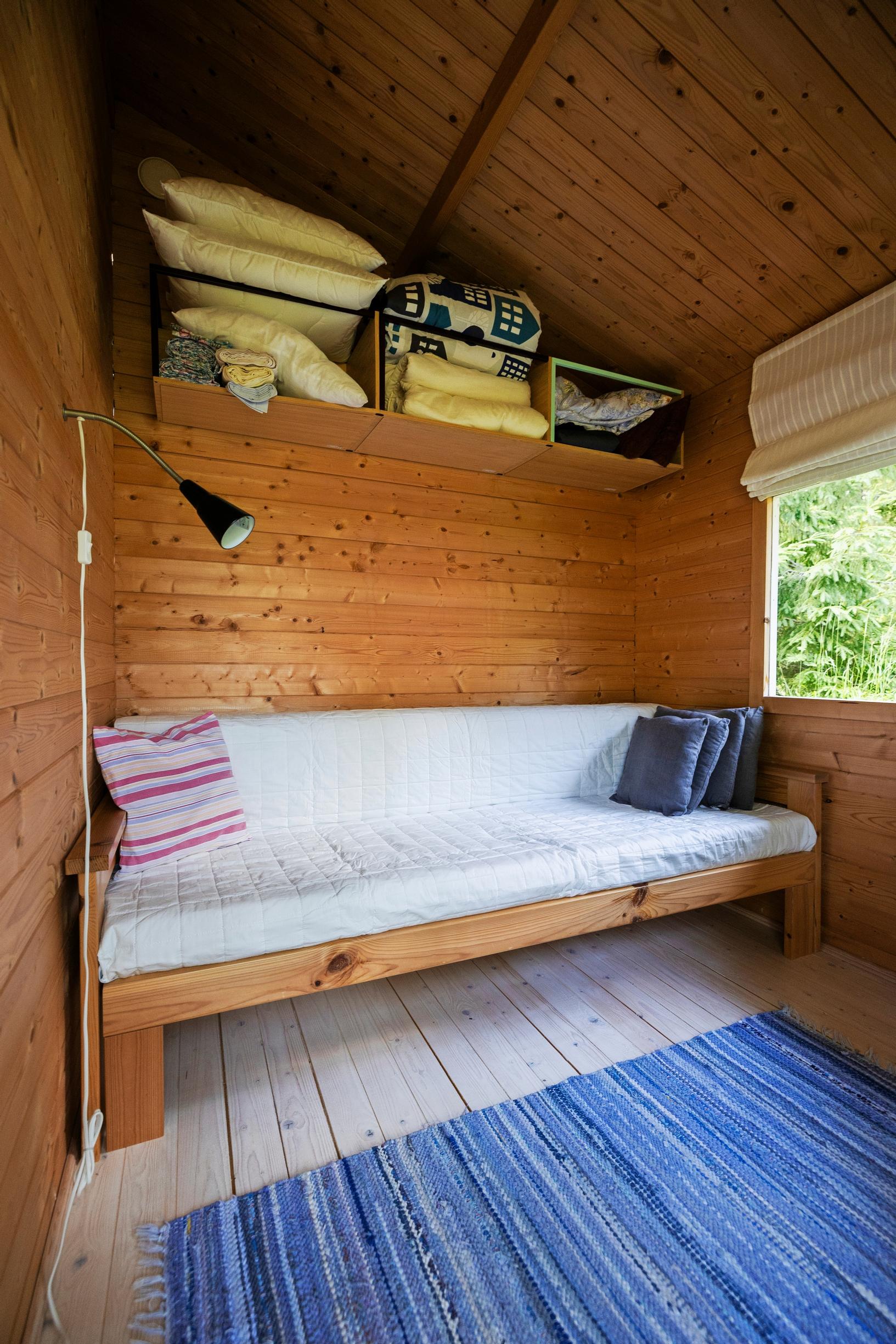 a wooden sofa with light cushions, textiles on a wooden shelf, wooden walls, and rag rugs
