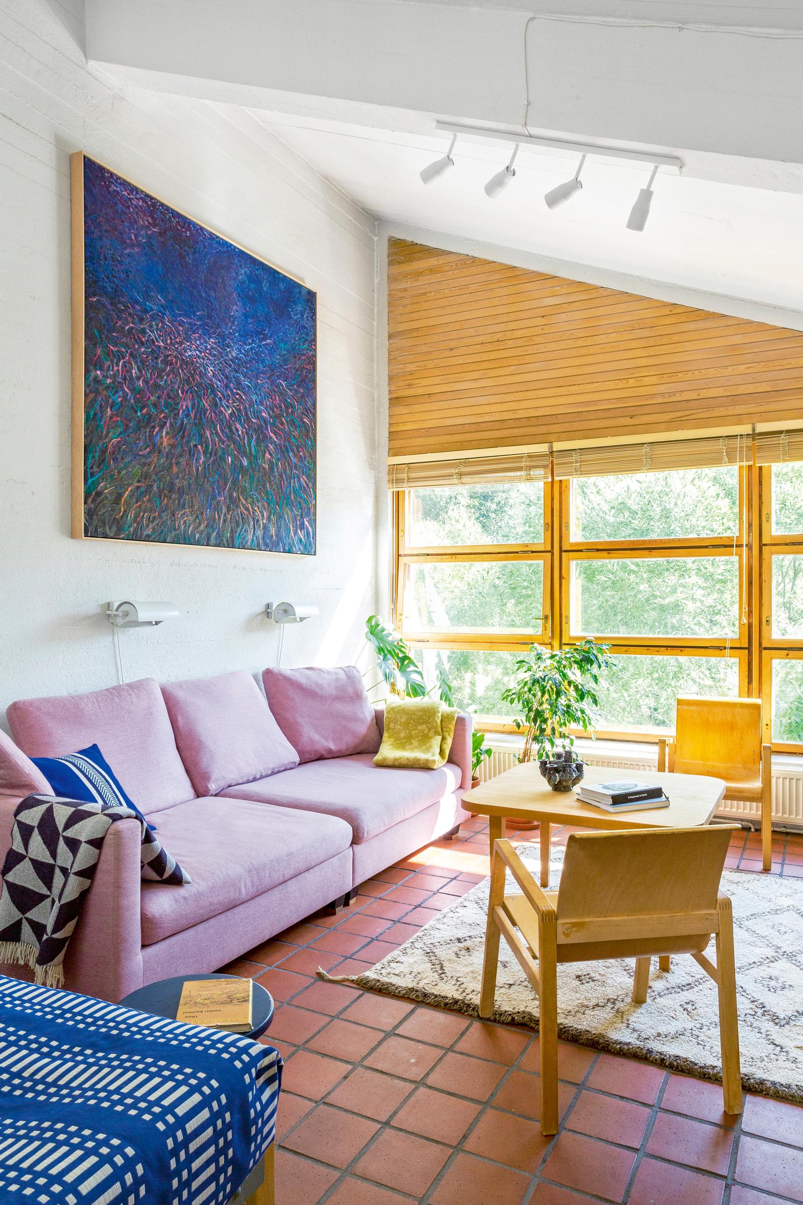 A pale lilac sofa, wooden chairs and table, a blue-toned painting, and terracotta-tiled floor