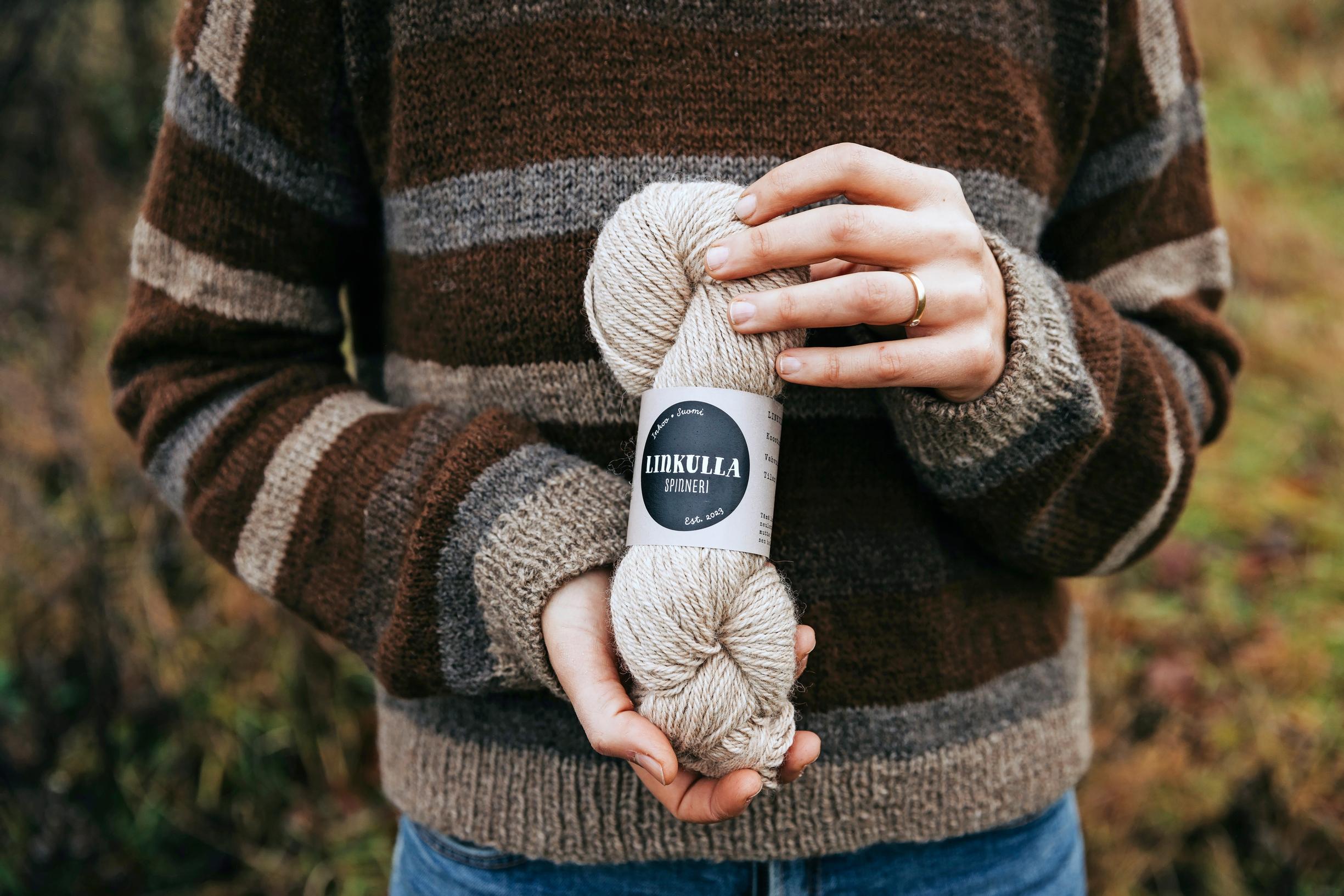 A person wearing a wool sweater holds a light-colored skein of yarn with a label