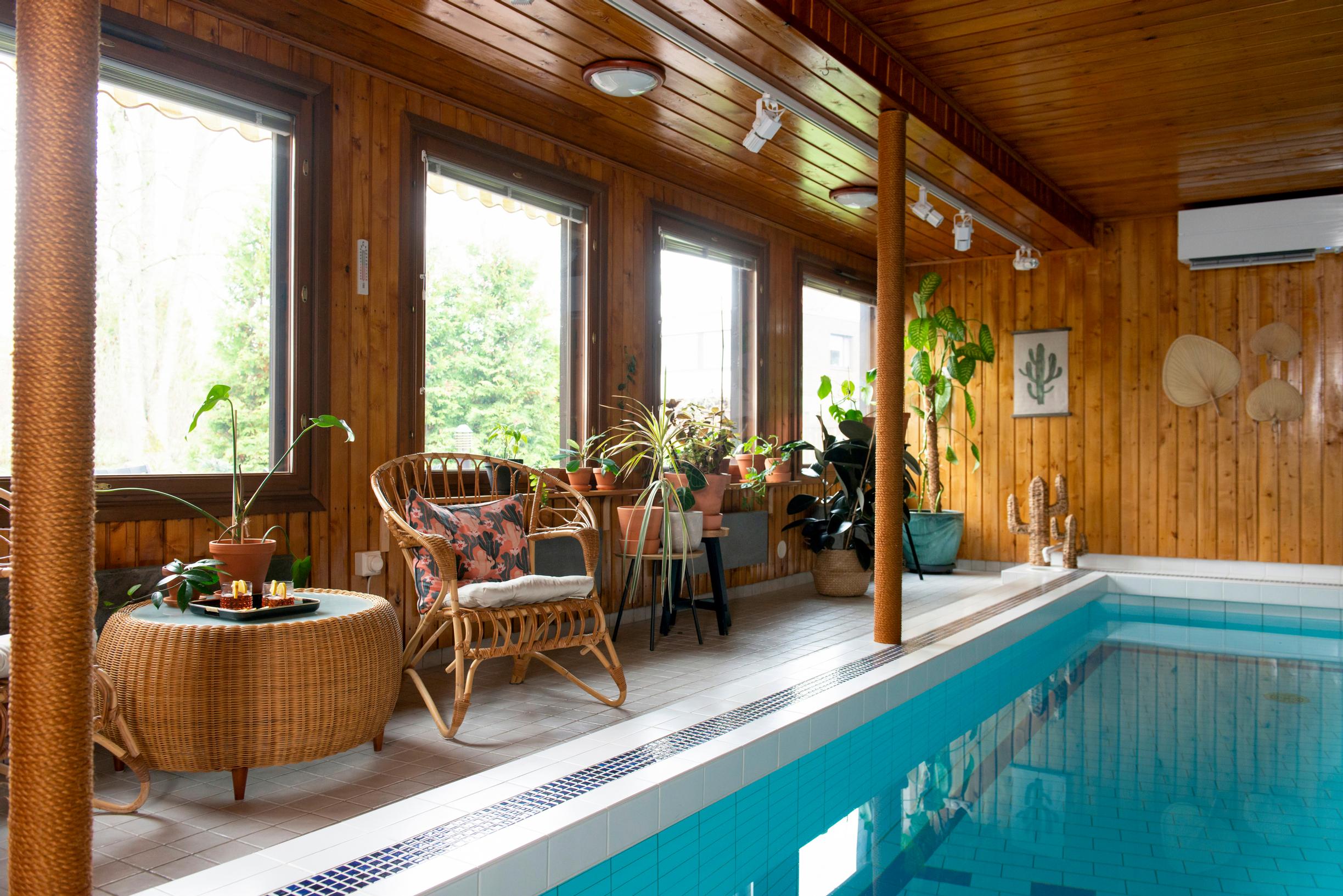 On the side wall is a row of large windows. The walls and ceiling are paneled. The floor surrounding the swimming pool is white tile.