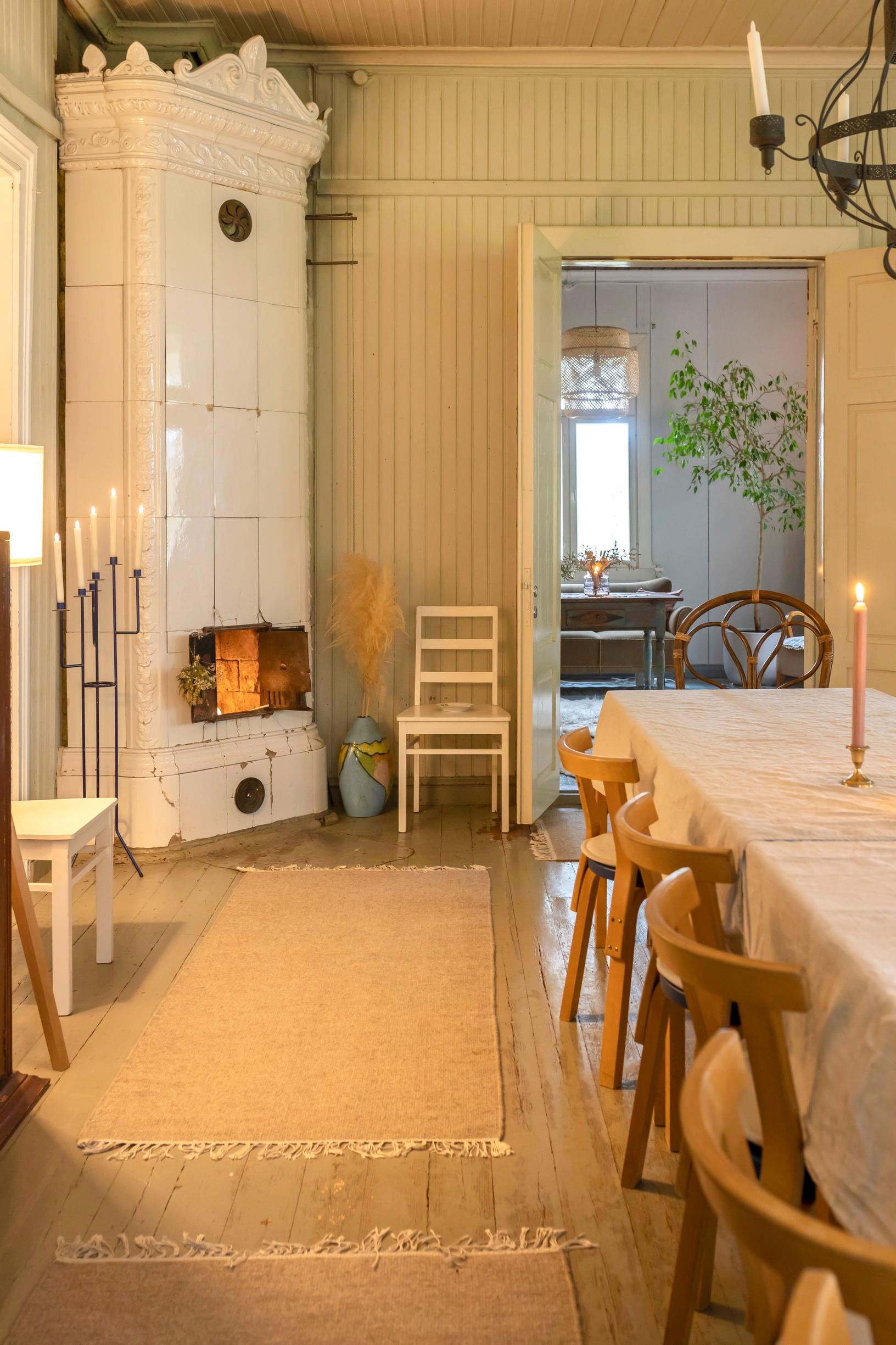 White tiled stove, long table, and light-colored rugs.