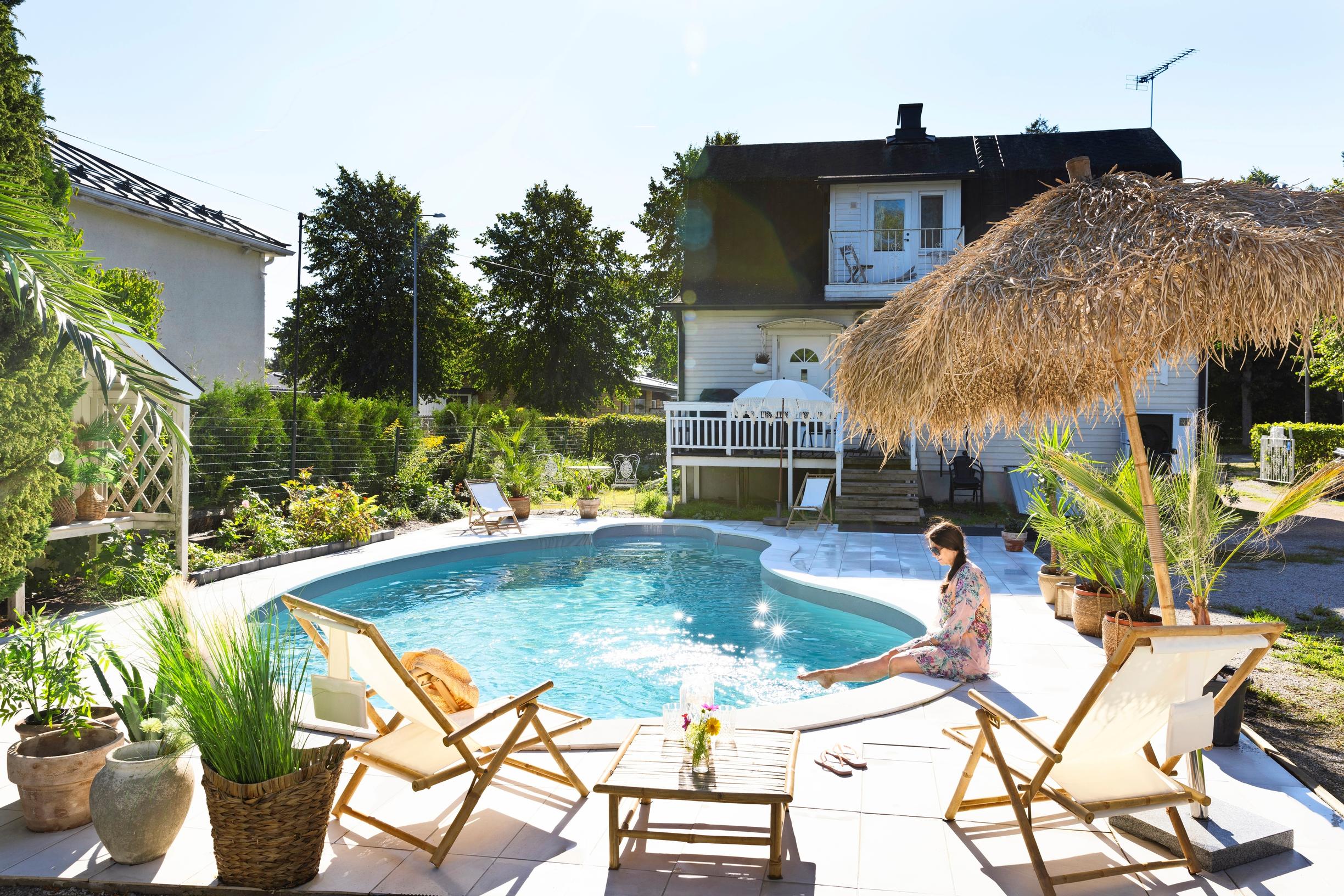 Marjaana Lundberg sits on the edge of her swimming pool, with an old house in the background and garden furniture and a sun umbrella in the foreground