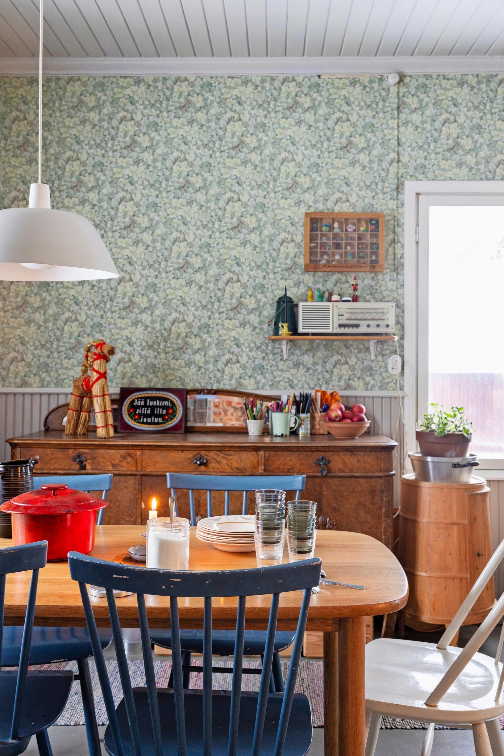 Colorful spindle-back chairs around the table, wallpaper and old birch chest of drawers, white ceiling lamp