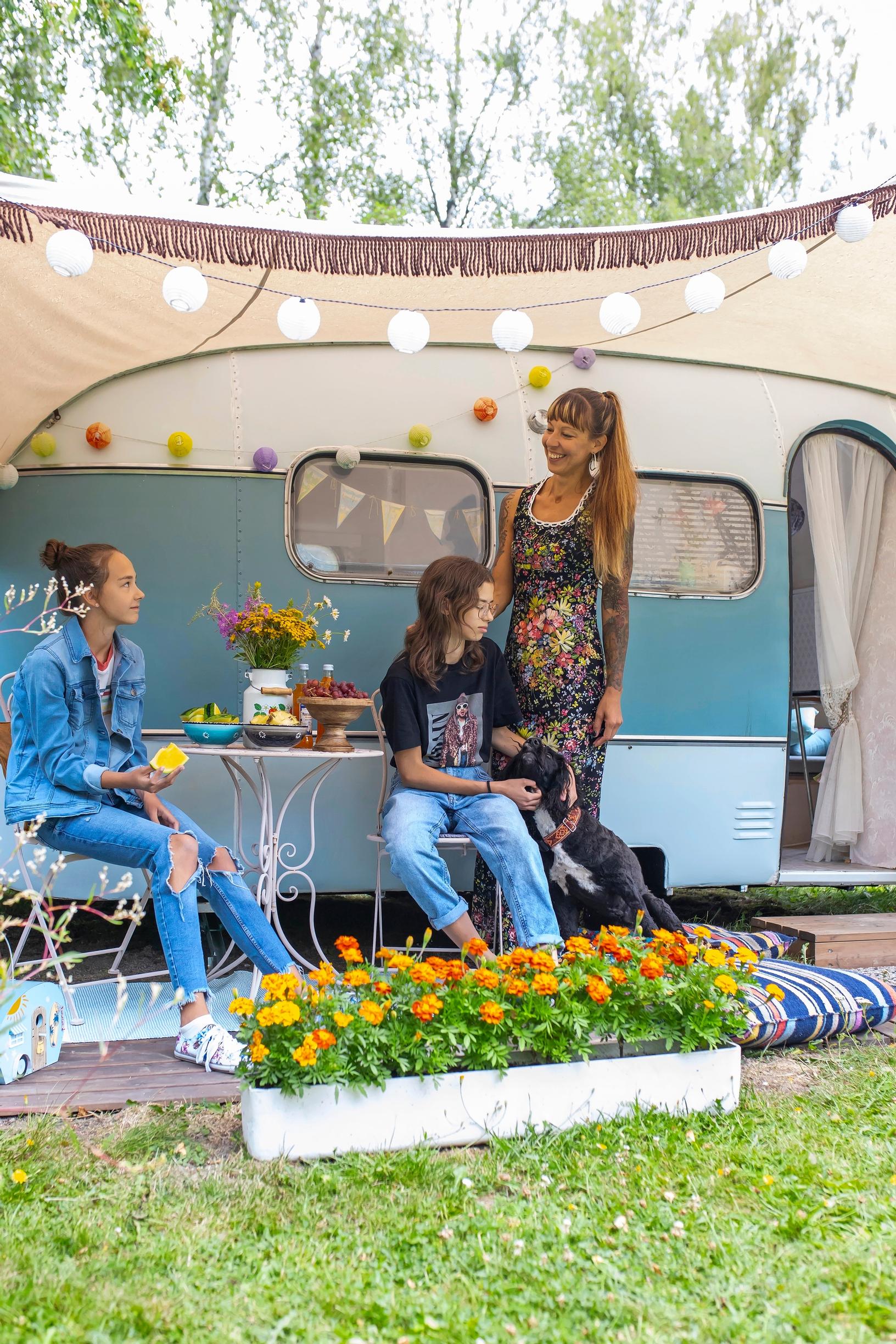 Two girls sitting and a mother standing beside an old caravan