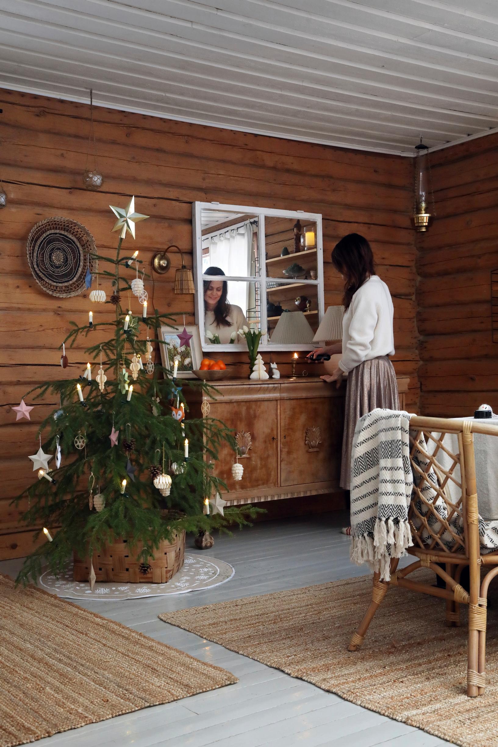 Ulla stands by the sideboard with a Christmas tree; on the wall is a window-like mirror, and there are brown rugs on the wooden floor.