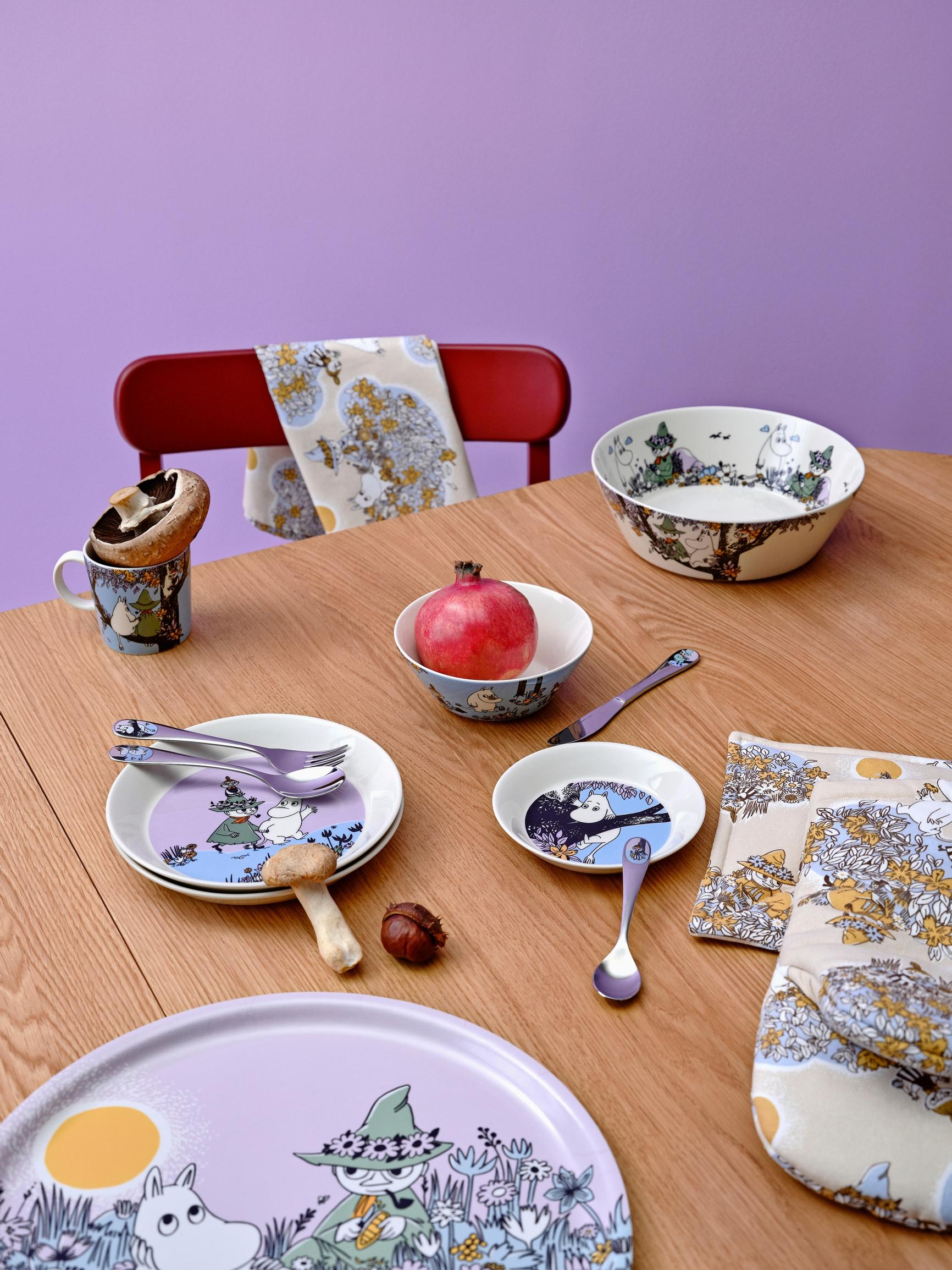 Plates in shades of lilac and blue, a mug, bowls, and oven mitts and pot holders featuring Moomin illustrations