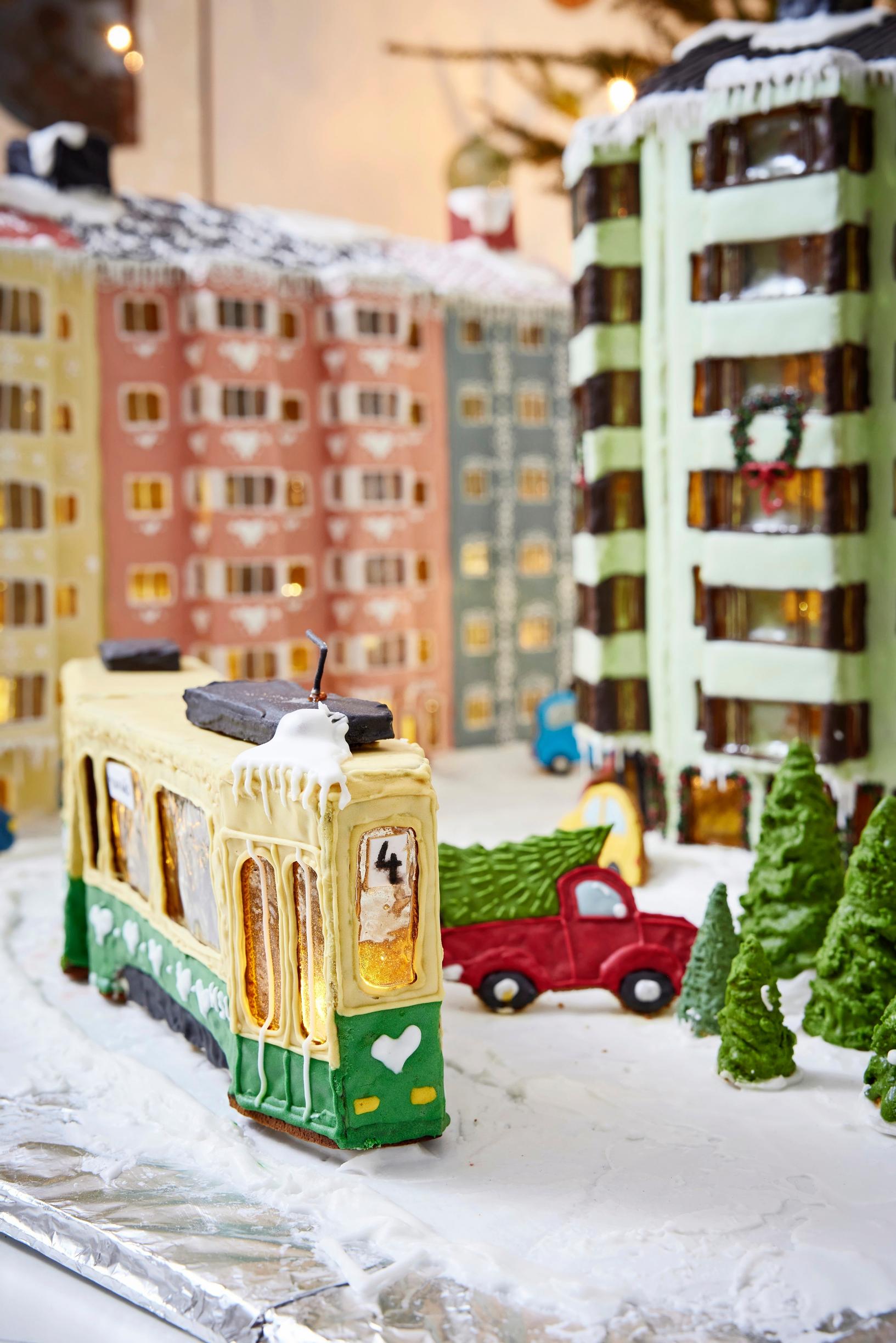 A gingerbread city block featuring a 1950s apartment building with bay windows covered in green and pink royal icing. A streetcar is in the middle.
