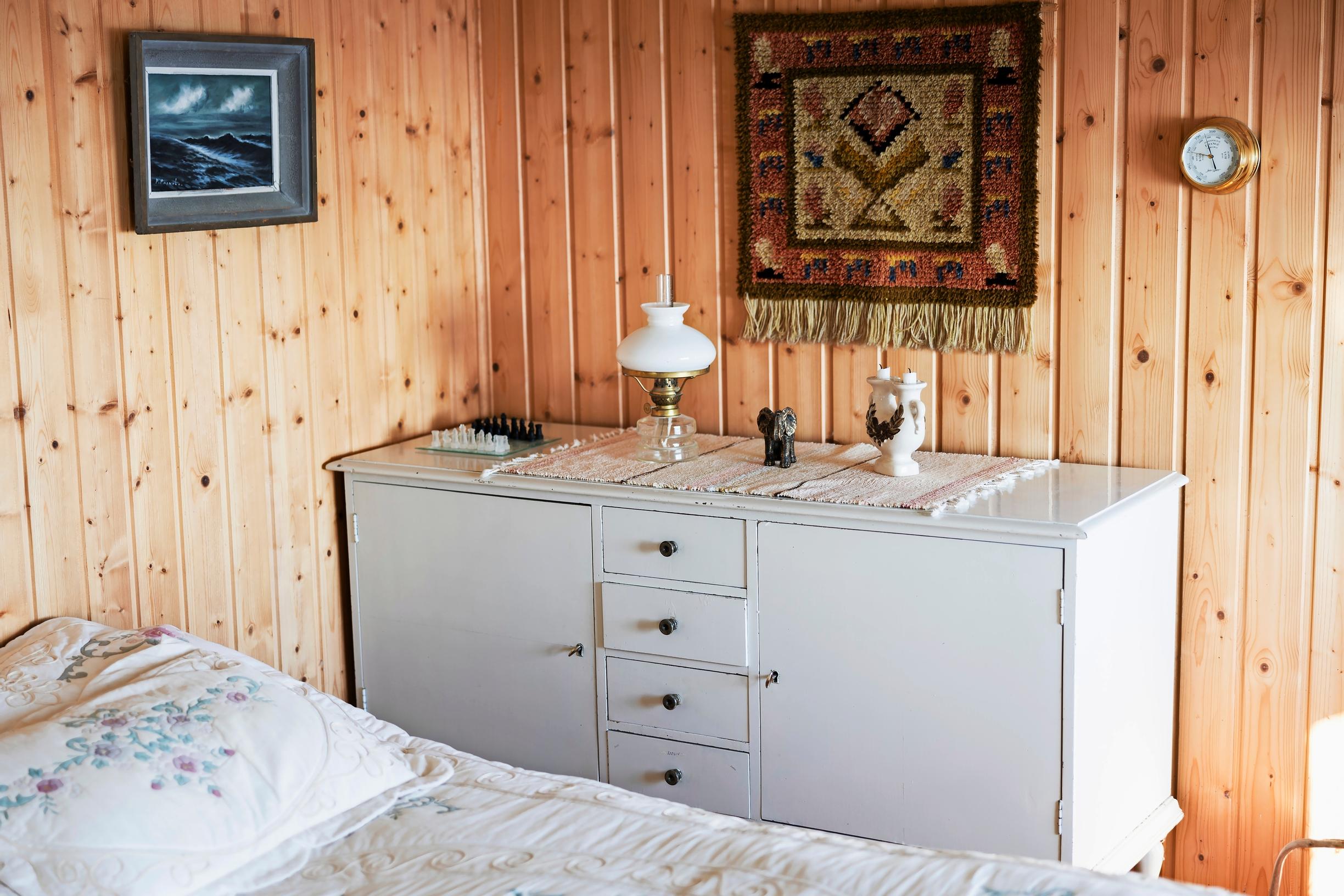 White dresser, bed, plank wall, oil lamp, and wall art including a tapestry and barometer
