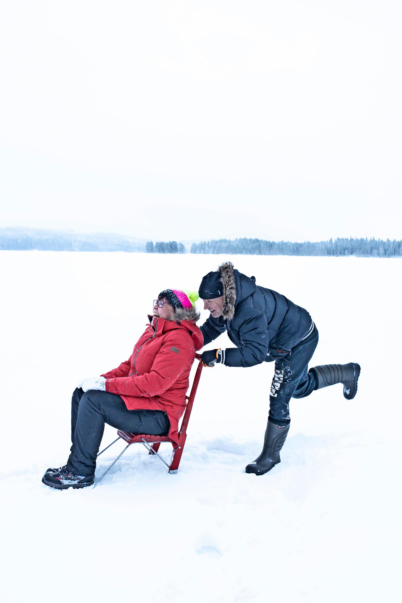A woman sits on a kicksled on the frozen lake while a man stands behind, pushing.