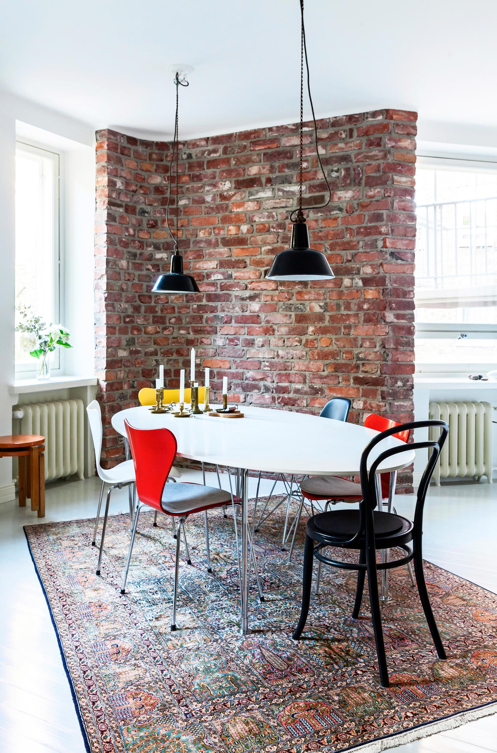 A white dining table with chairs in various colors stands before the brick corner. An oriental rug lies beneath.