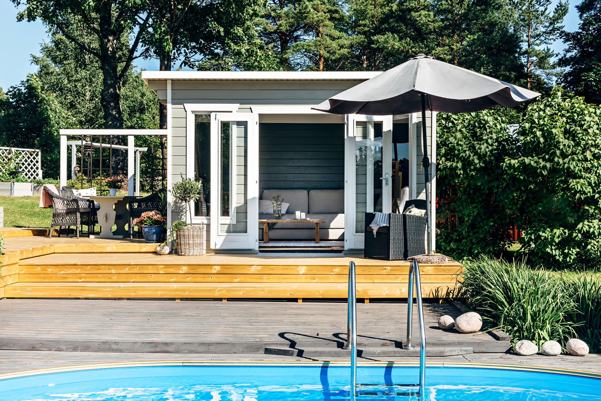 A gray outbuilding with a swimming pool in front of it, along with garden furniture, a terrace, and greenery around