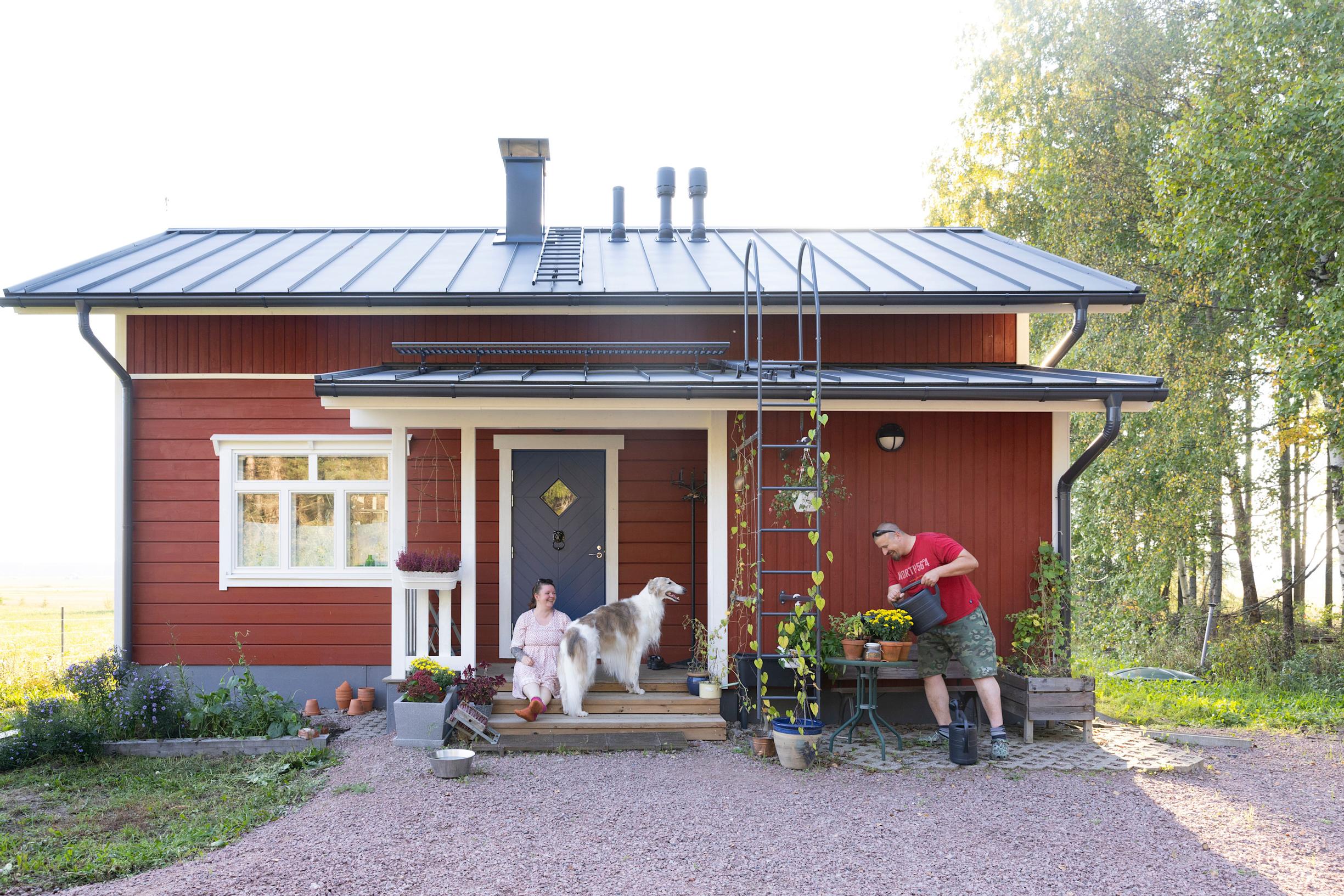 Tiia Inkinen stands on the terrace of her red house with her dog while her partner waters flowers.