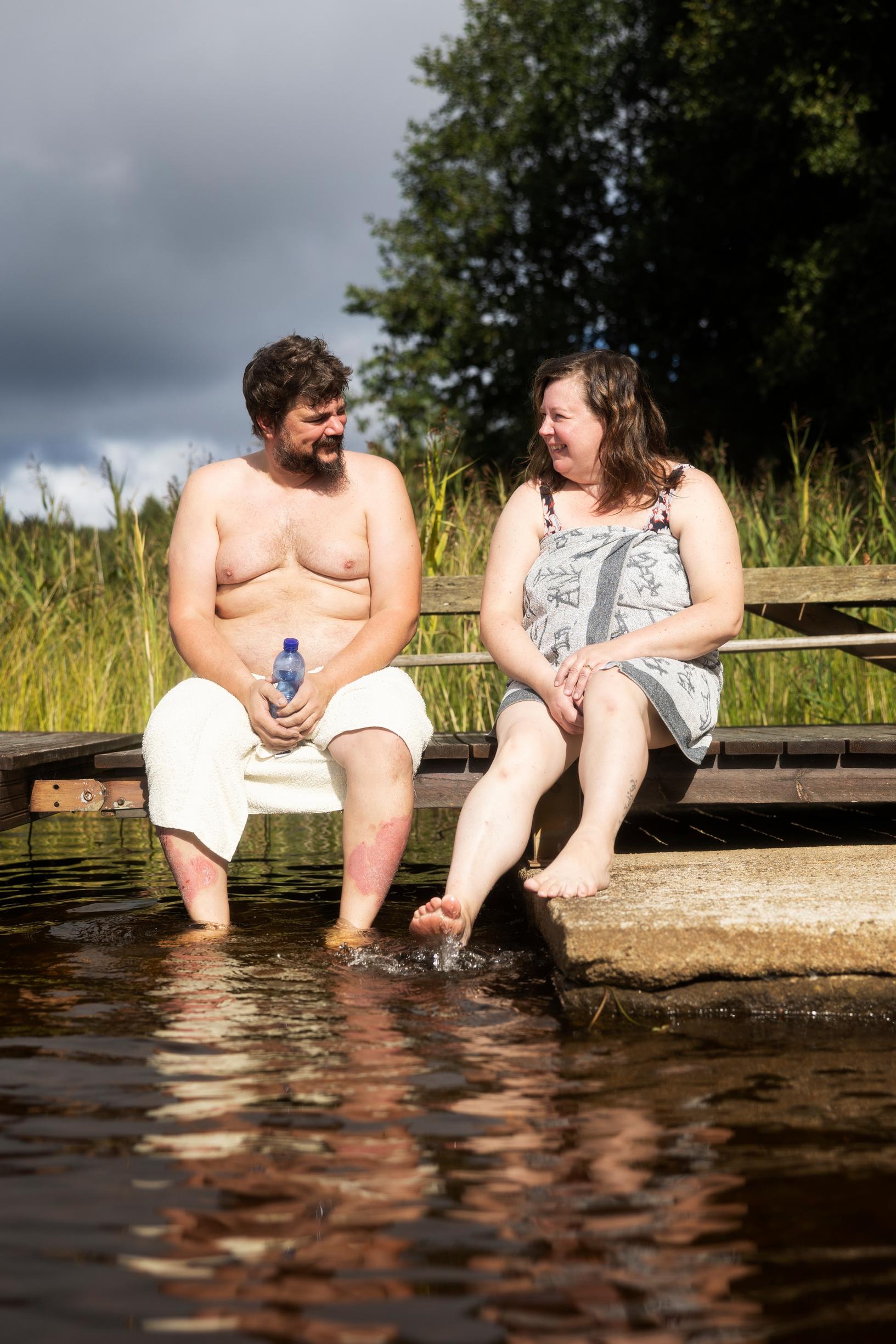 A man and a woman sitting on a dock, looking at each other. The man is holding a bottle, with reeds in the background.