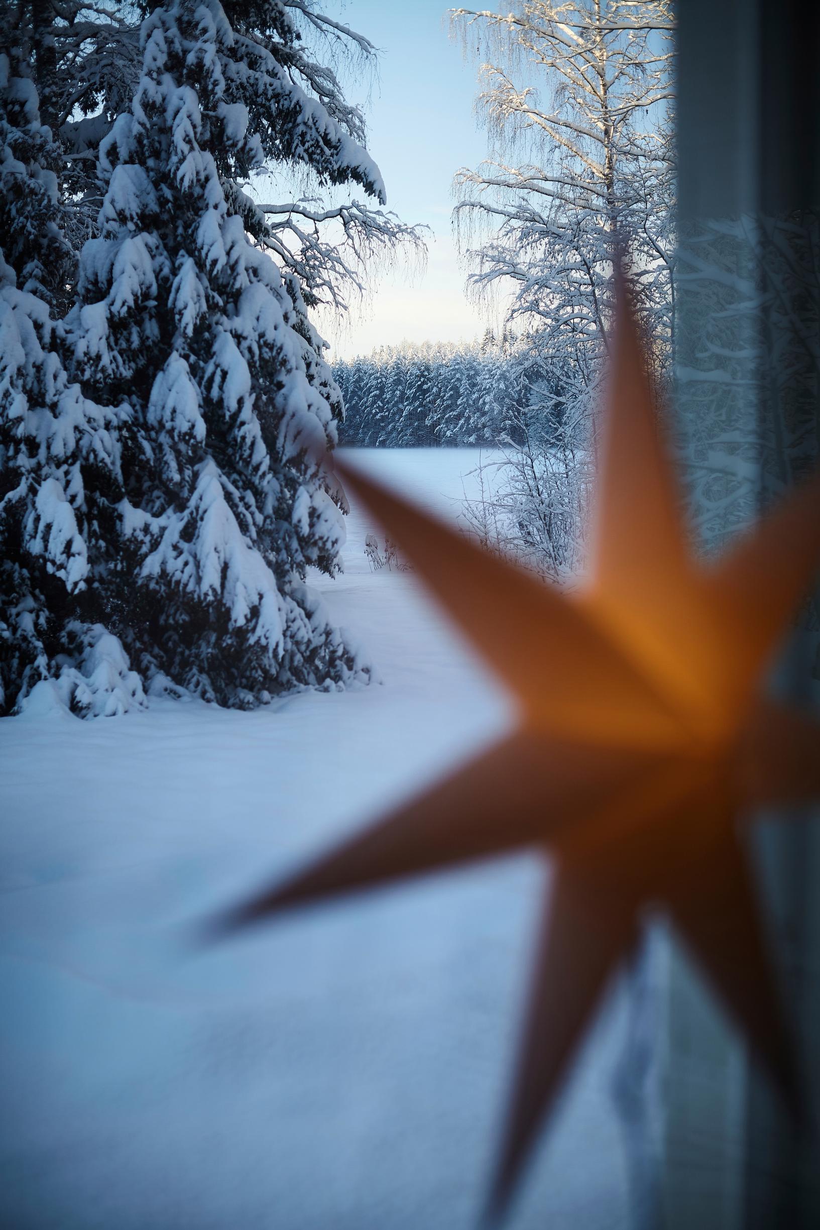 A light-colored Christmas star hanging from the window in the foreground, with a snowy lakeshore and a spruce tree in the background