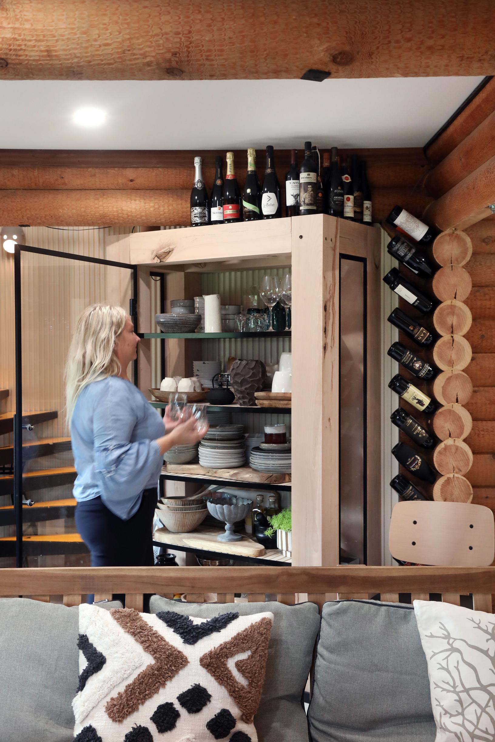 In the kitchen, a wooden china cabinet with a glass door. The log wall has holes drilled for wine bottles.