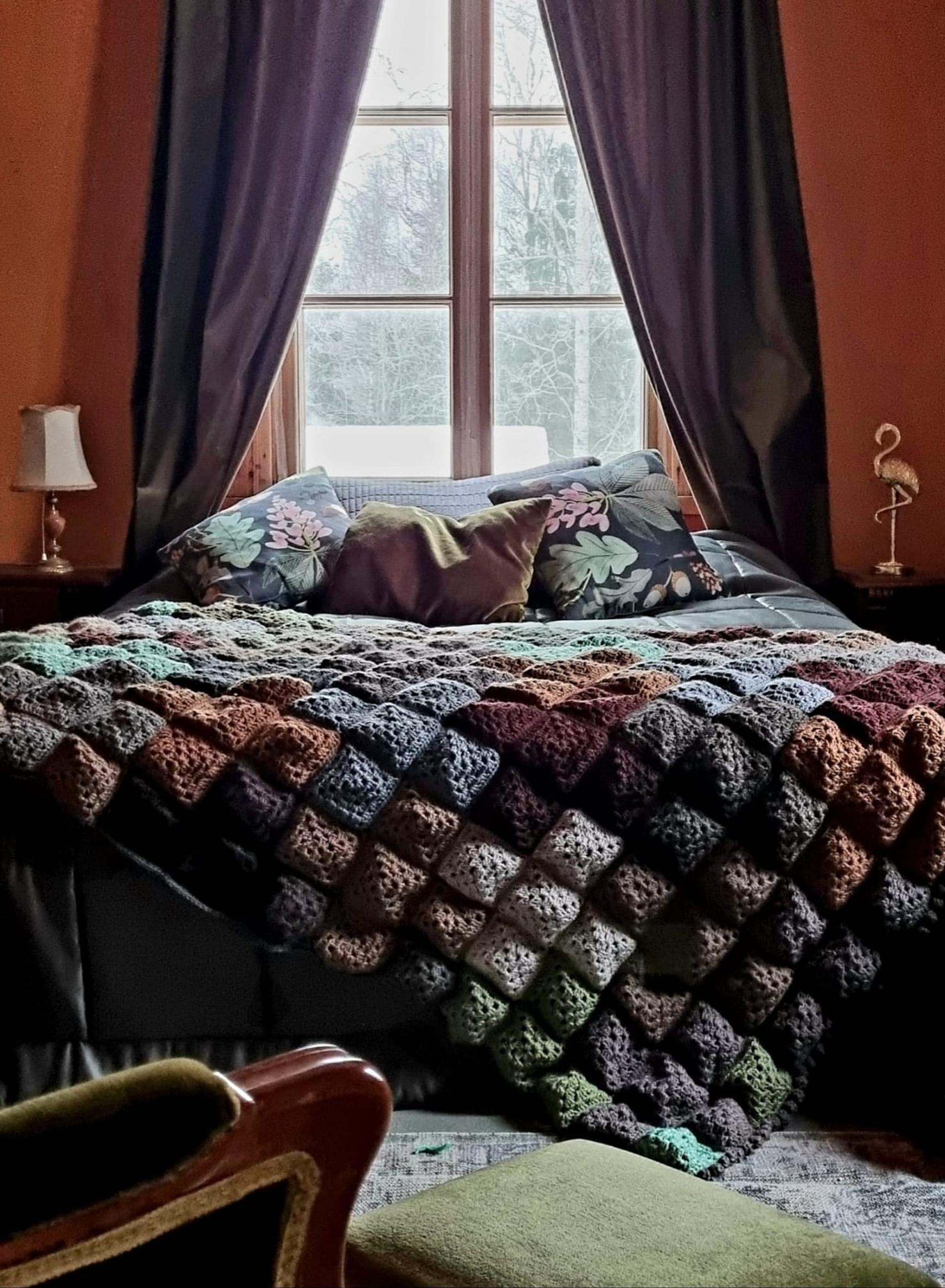 A greenish-brown crocheted bedspread on a double bed, with a window, curtains, and table lamps on bedside tables in the background