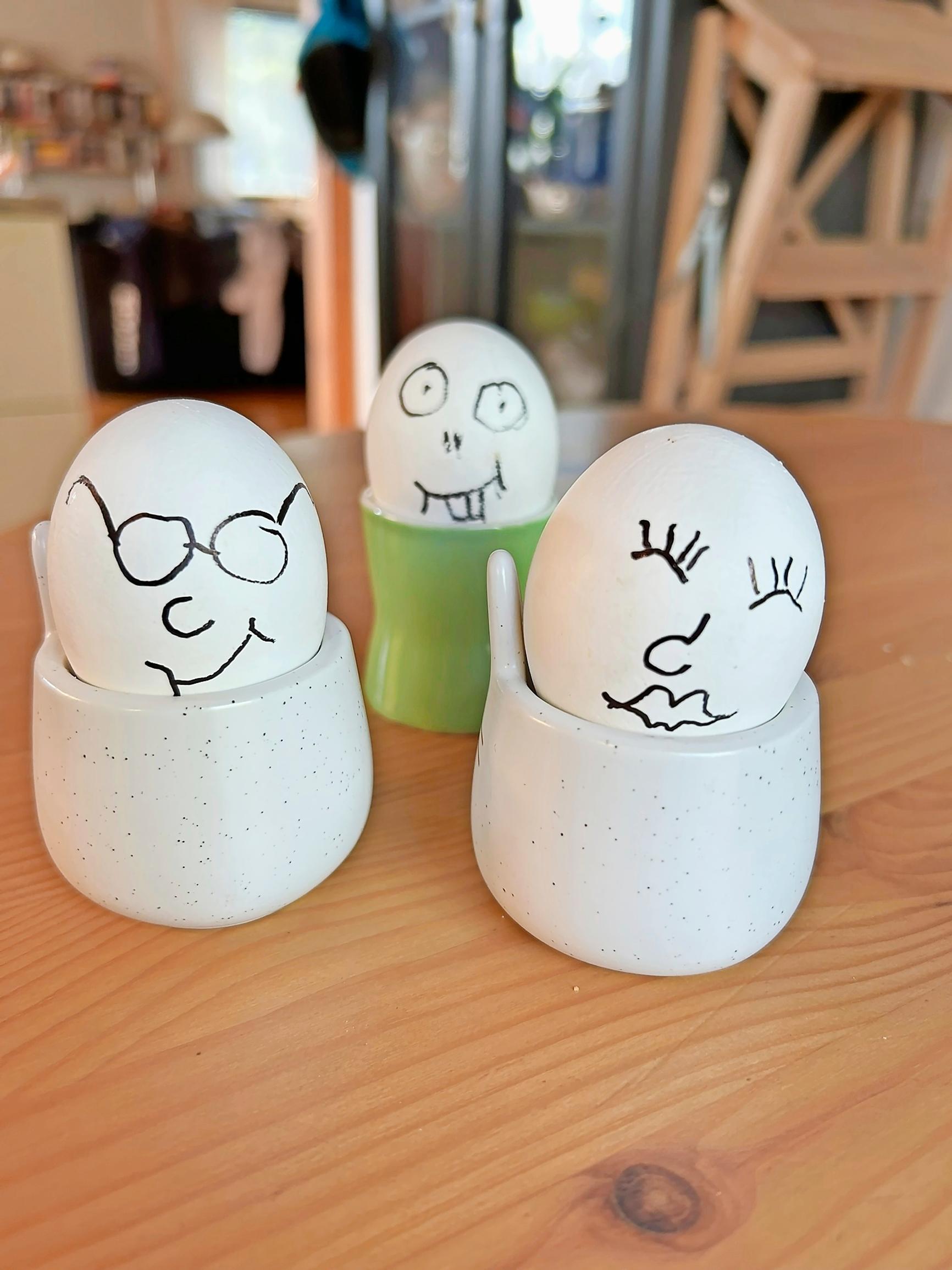 Three eggs in cups with faces drawn on them in black marker.