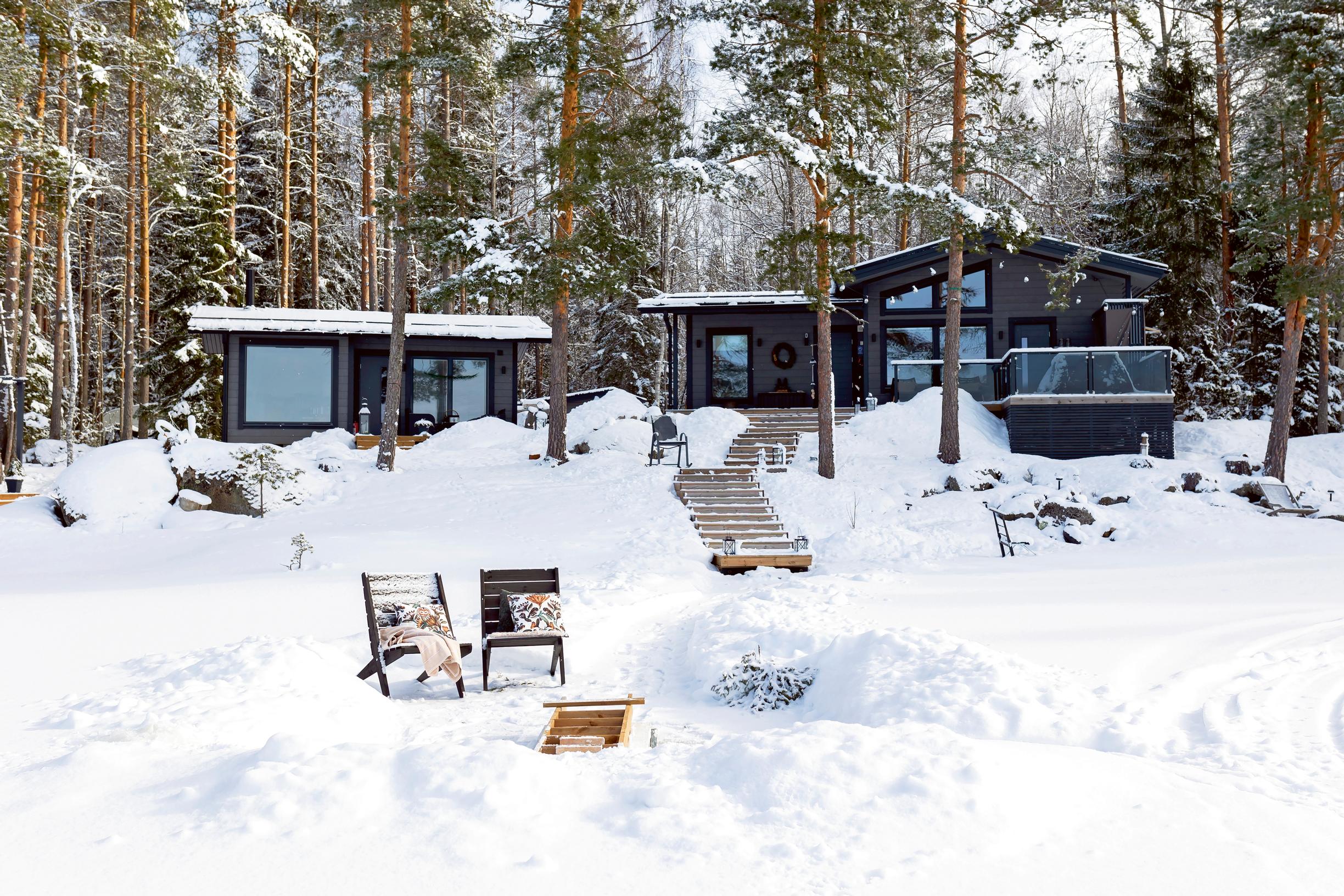 A black cottage and lakeside sauna set in a snowy landscape by the lake, with two sun loungers on the shore