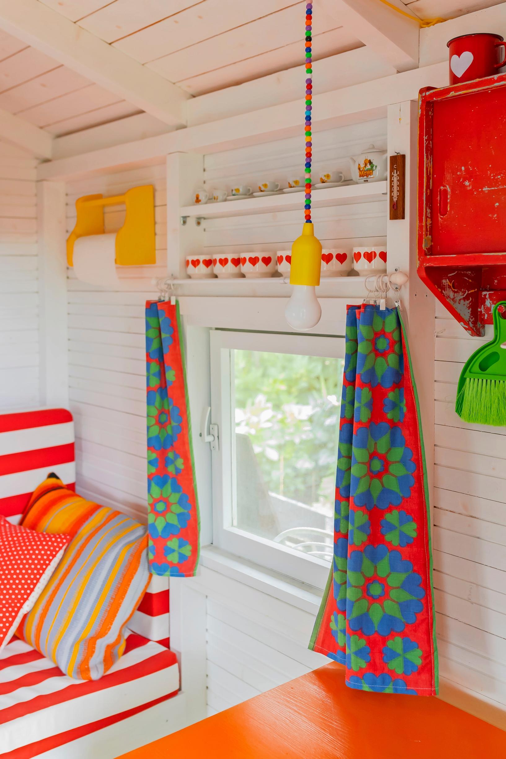 red, blue, and green retro curtains, a white wooden wall, striped cushions on the sofa, and red-and-white dishes on the shelf