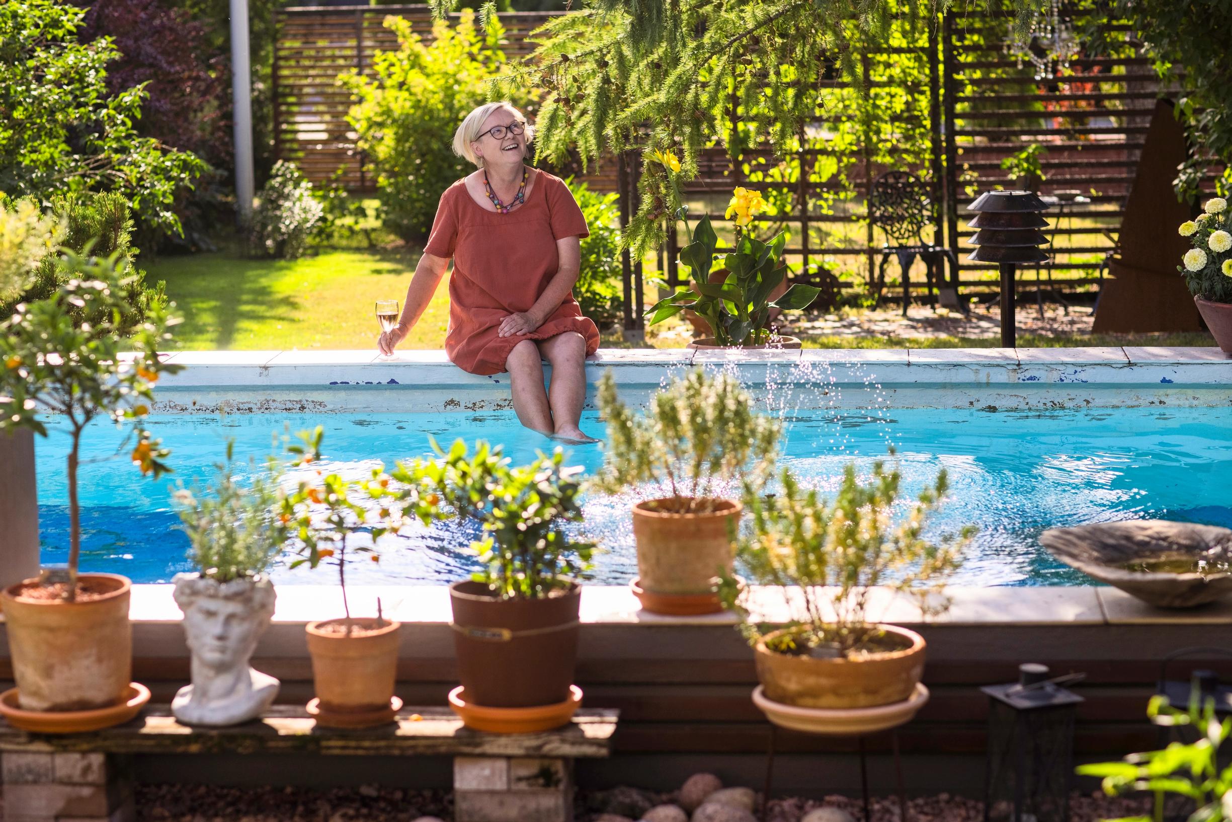 Merja Heinonen sits on the edge of the swimming pool, with potted plants in the foreground