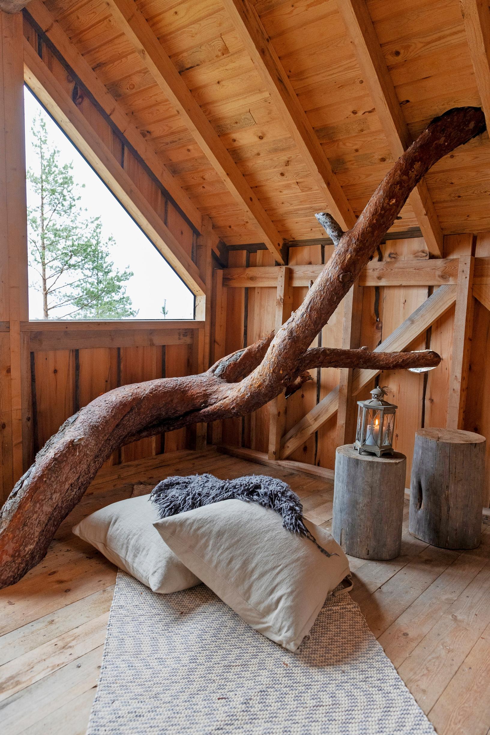 A thick tree trunk running through a wooden treehouse with a window, logs, a lantern inside, and pillows plus a rug on the floor