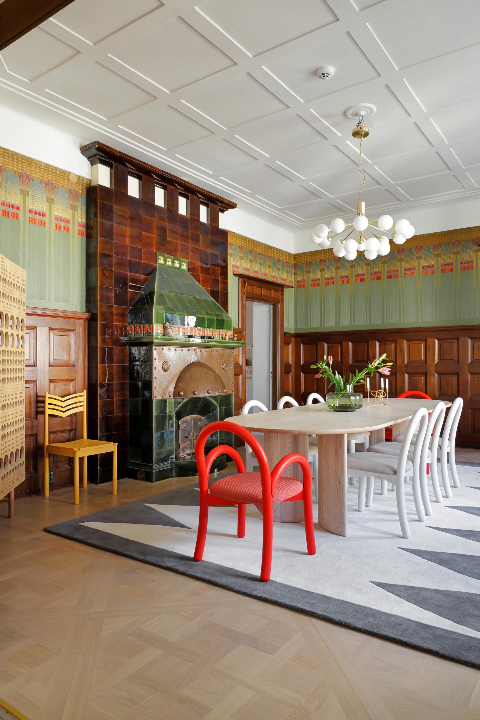multi-colored national romantic, low tile stove, a long table with chairs, colorful walls, and a white ceiling lamp