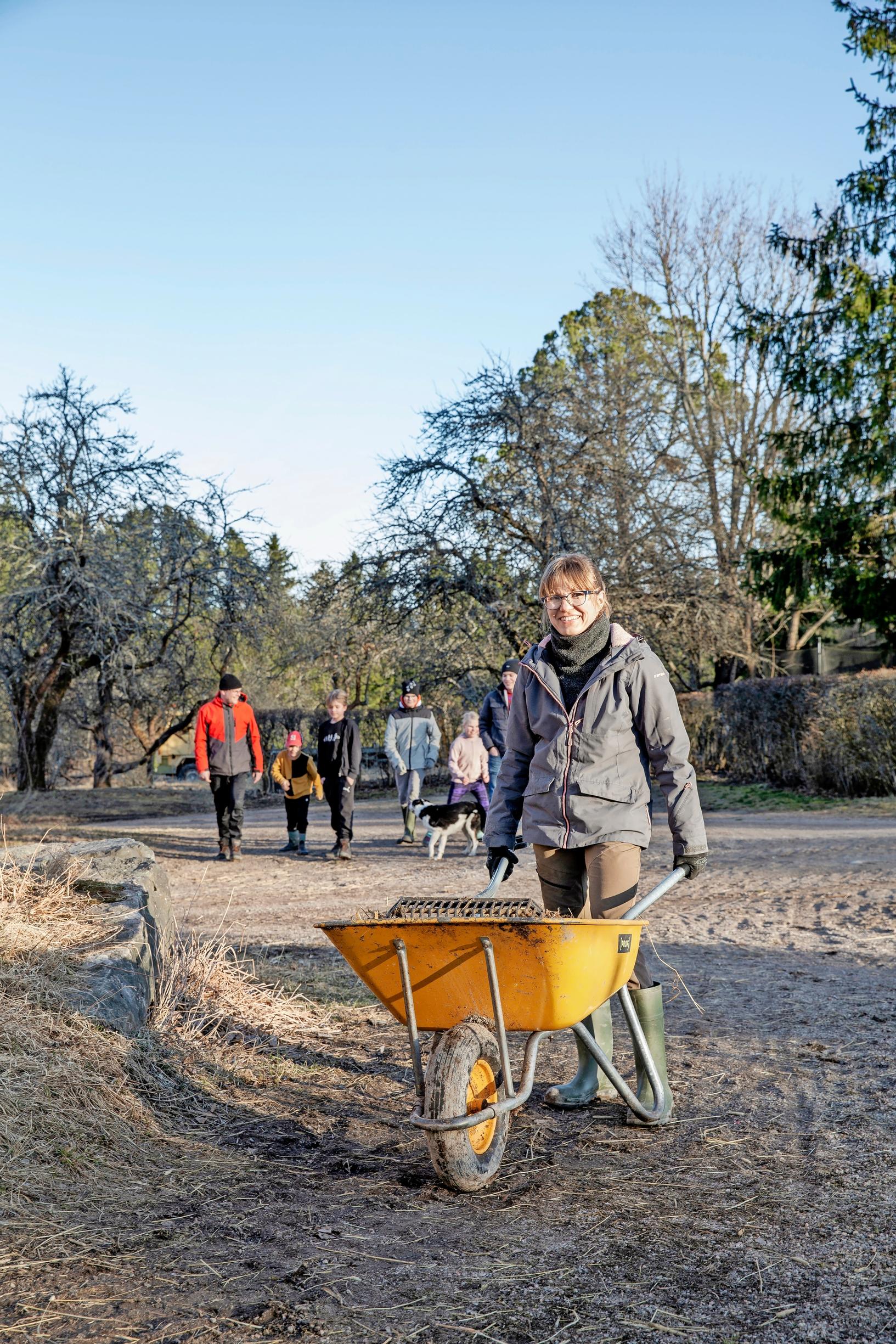 A woman pushing a wheelbarrow on a gravel road, her family visible in the background
