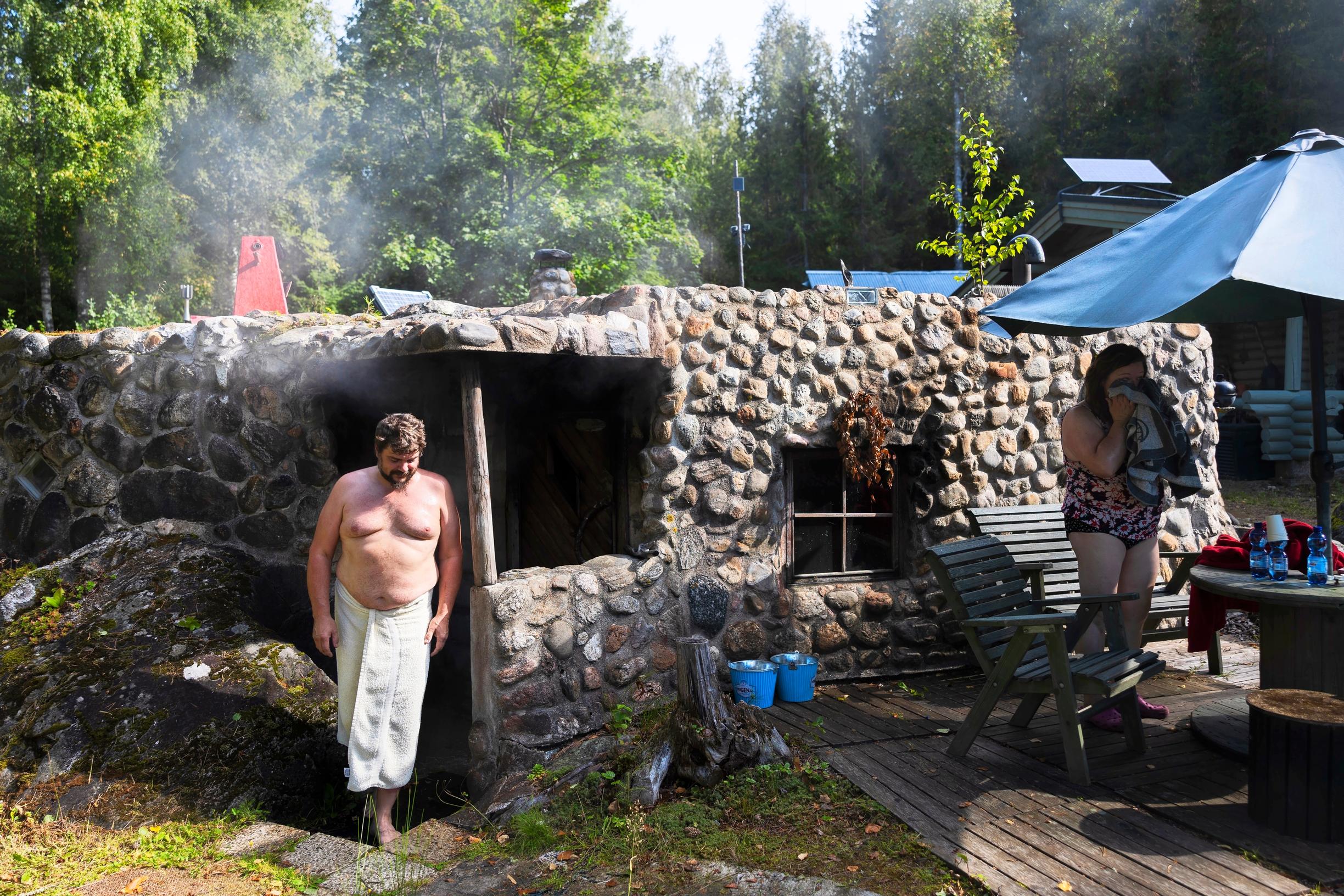 A man and a woman cooling off in front of a natural-stone smoke sauna, with a garden set on the right.
