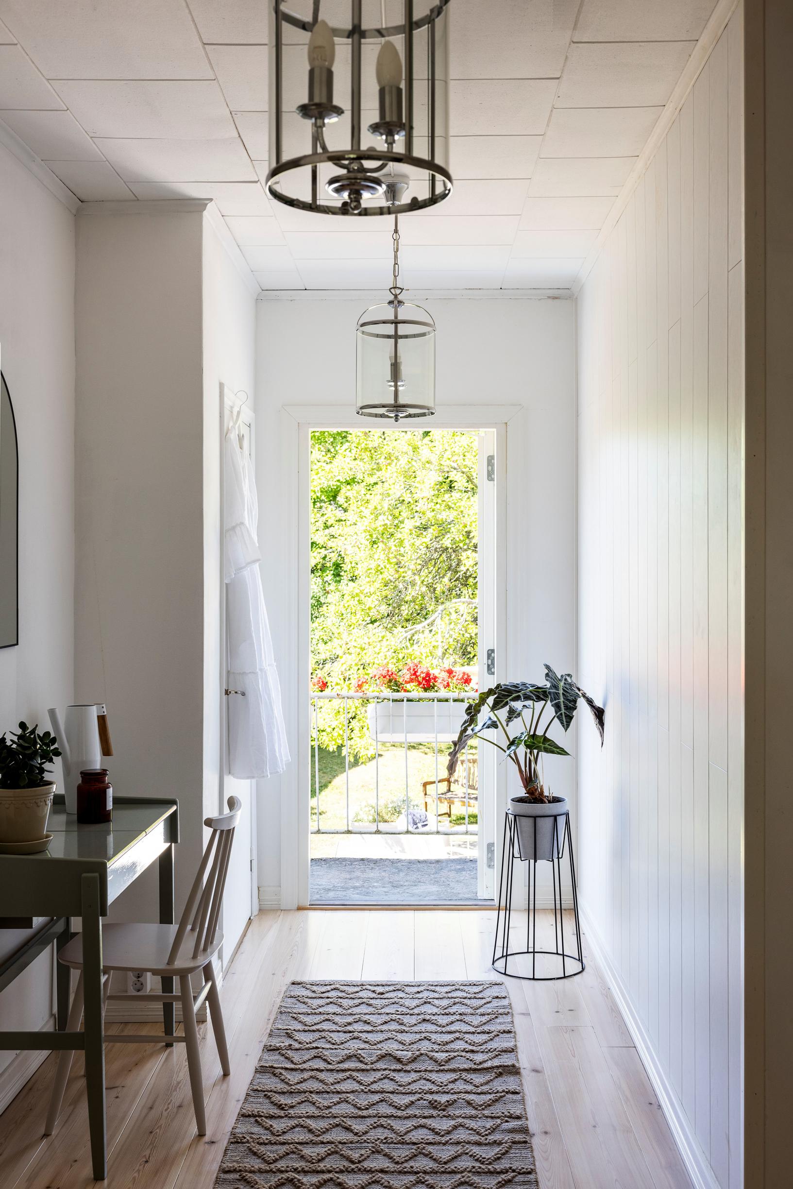In the foreground is a desk and chair, and at the end of the hallway the balcony provides a garden view