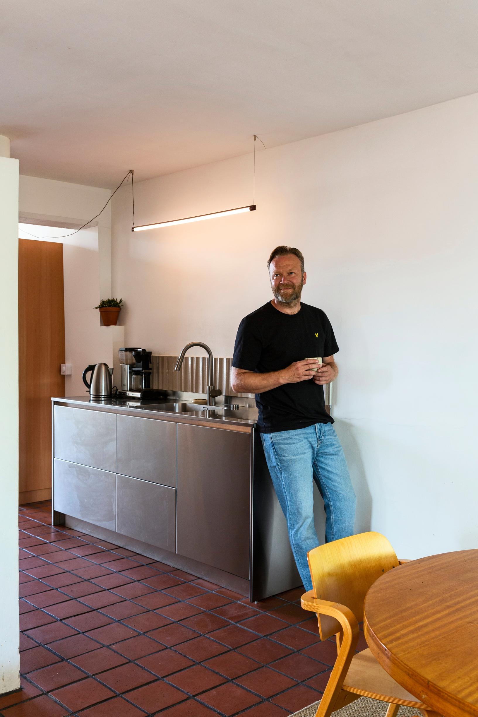 Eemil Karila standing in his home’s kitchen, leaning against the cabinet; in the foreground a table and chair, tiled floor