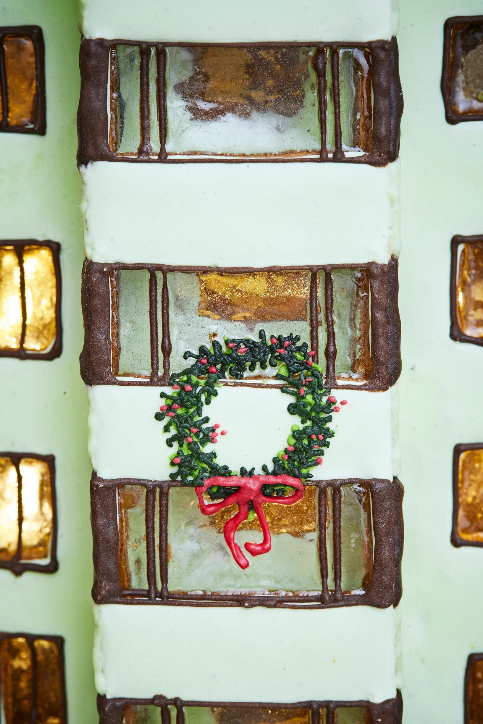 A gingerbread city block featuring a 1950s apartment building with bay windows covered in green royal icing. A Christmas wreath hangs from one of the bay windows.