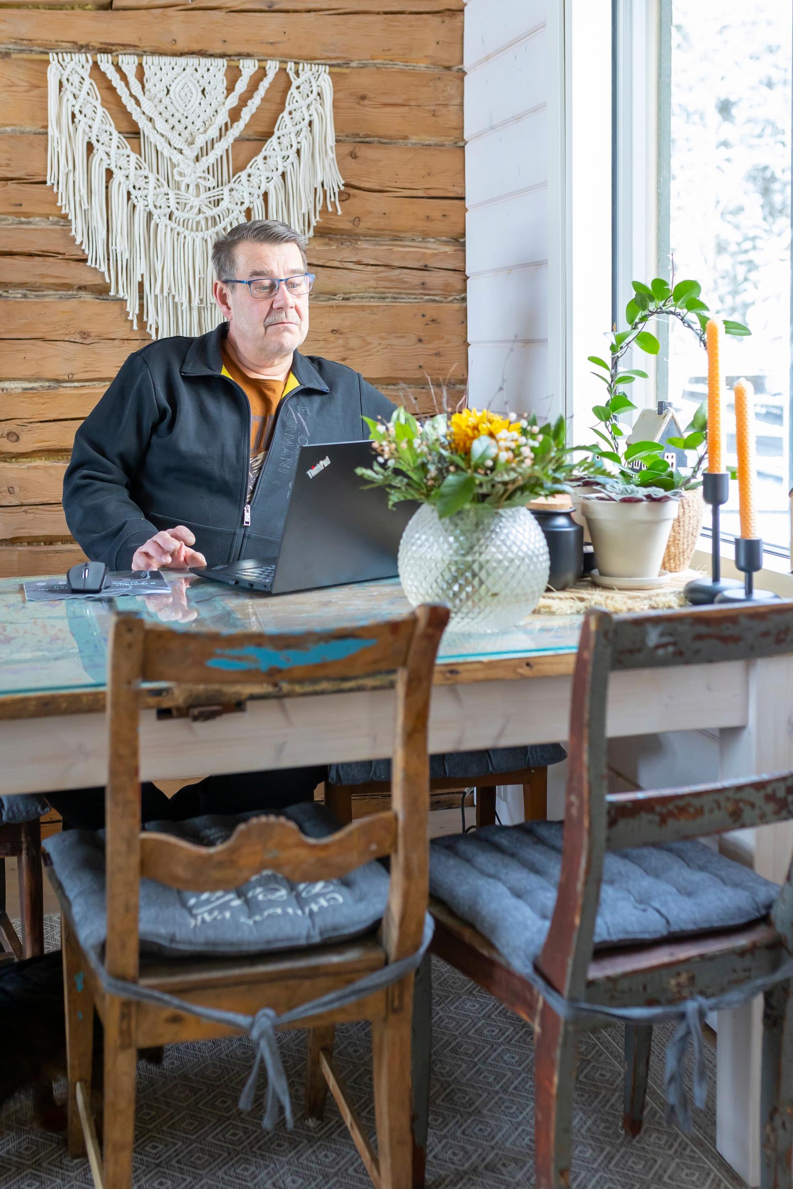 A man sits at a table looking at his laptop; two chairs, a houseplant, a flower vase, and a window