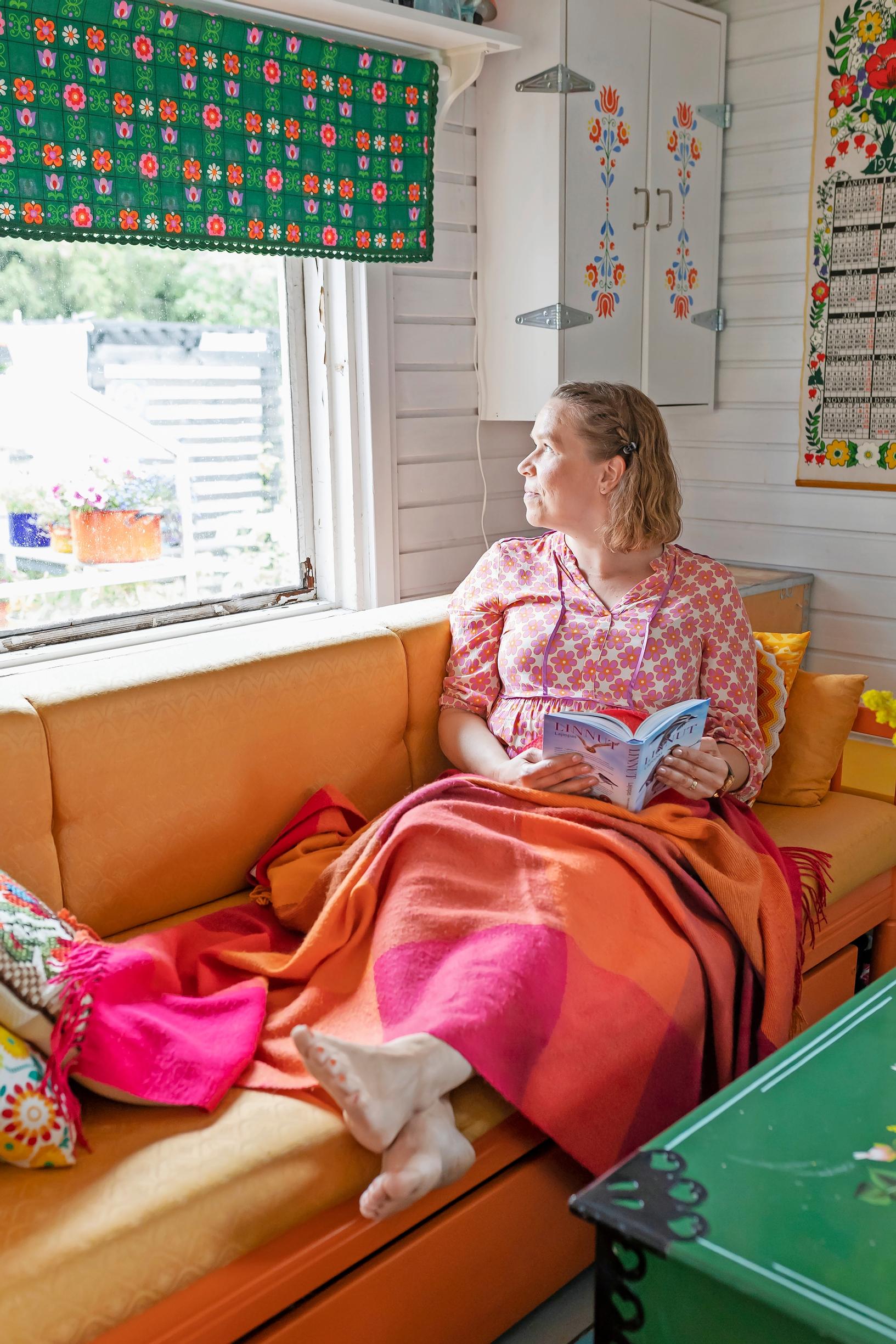 Leni Joutsjoki sitting on an orange sofa with a book in hand, looking outside